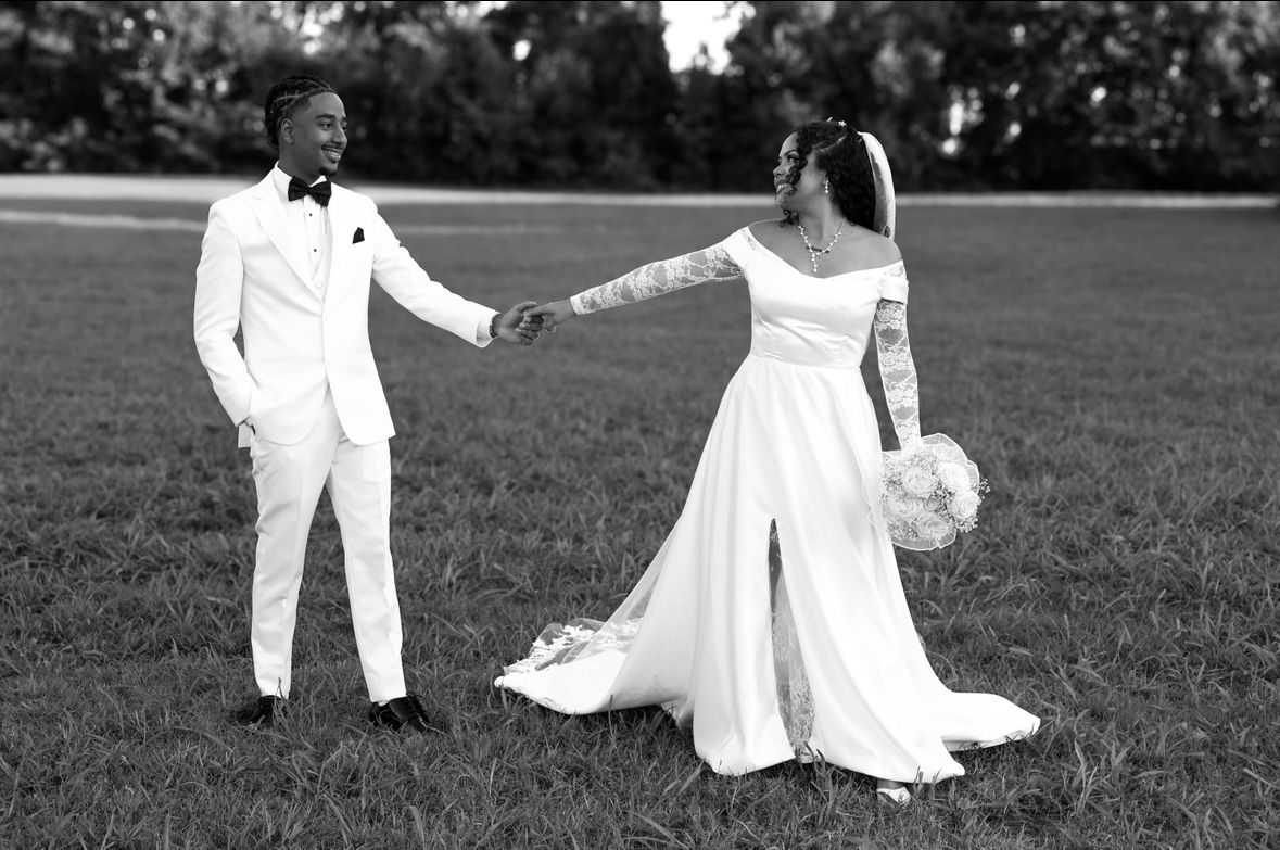 Black and white photo of a couple in wedding attire holding hands in a green field.