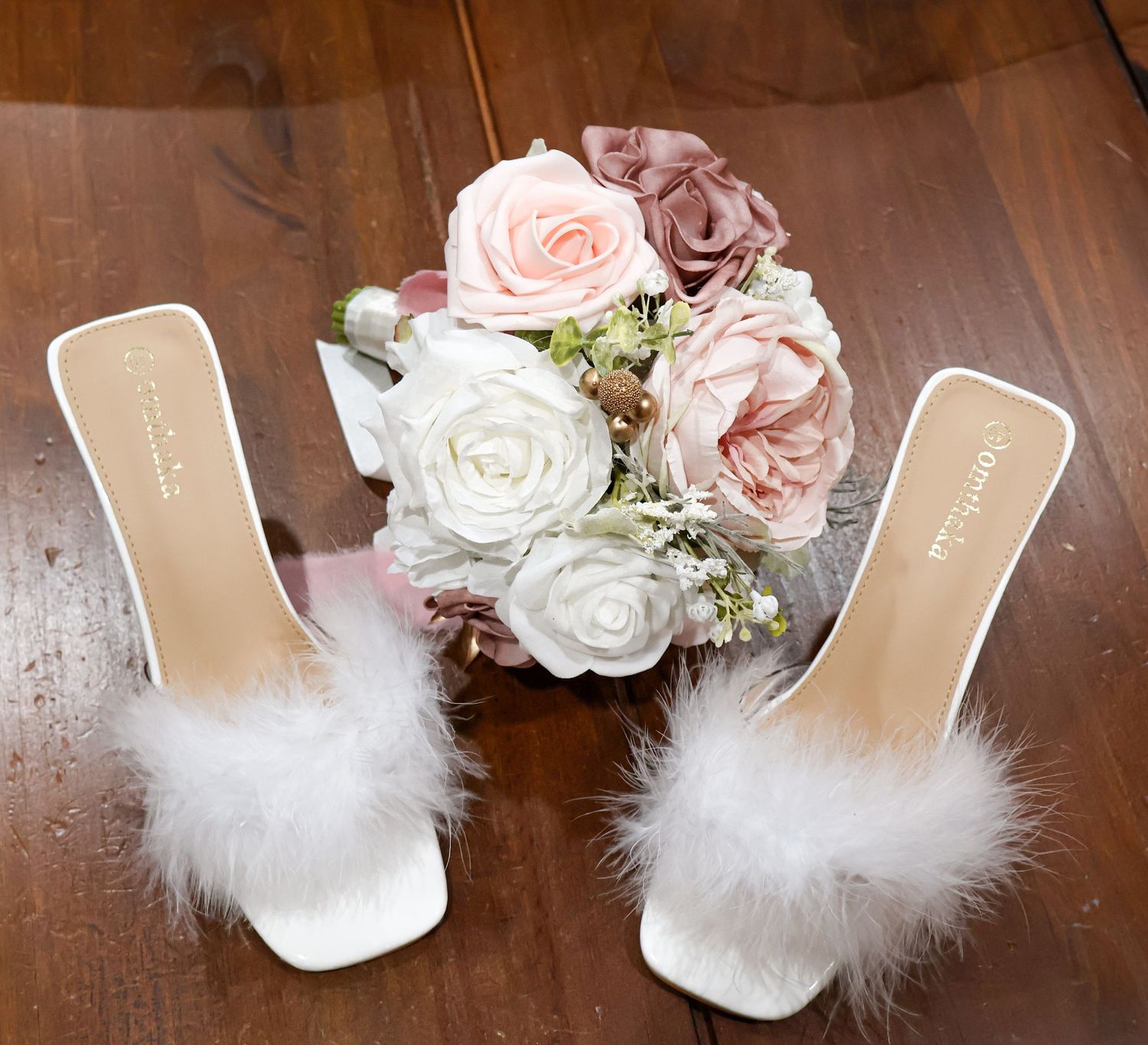 White feather bridal heels beside pink and white rose wedding bouquet on wooden surface.