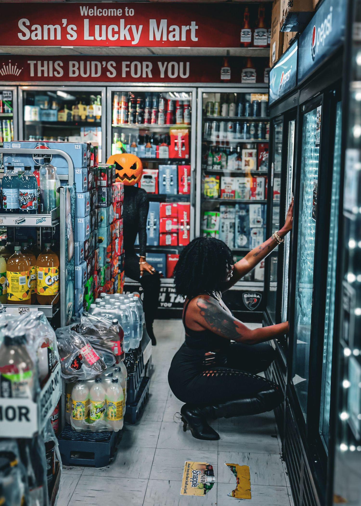 A woman crouches by a refrigerator in Sam's Lucky Mart convenience store with Halloween pumpkin decor.