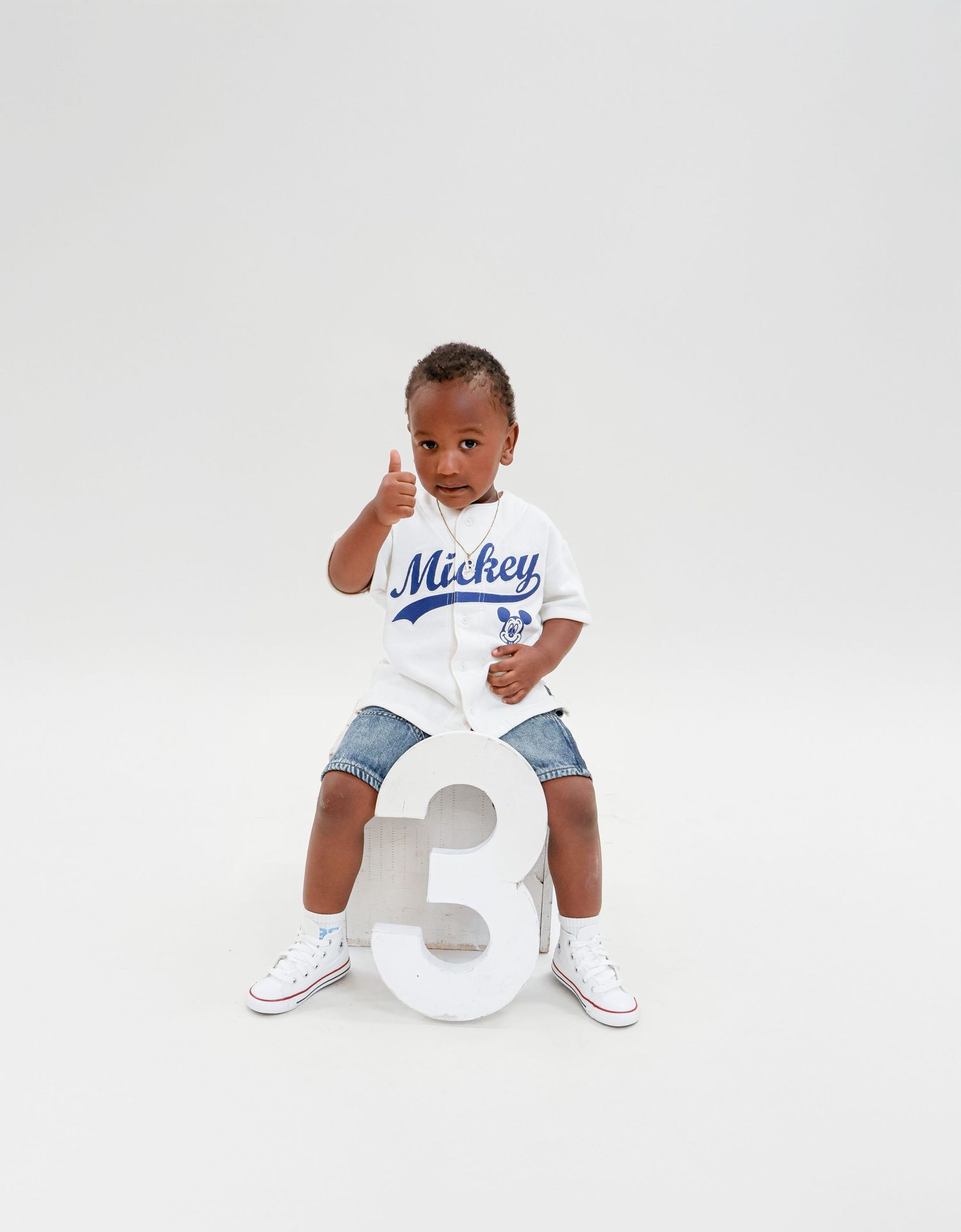 A toddler in a Mickey Mouse jersey gives a thumbs up while sitting on a large number three prop.