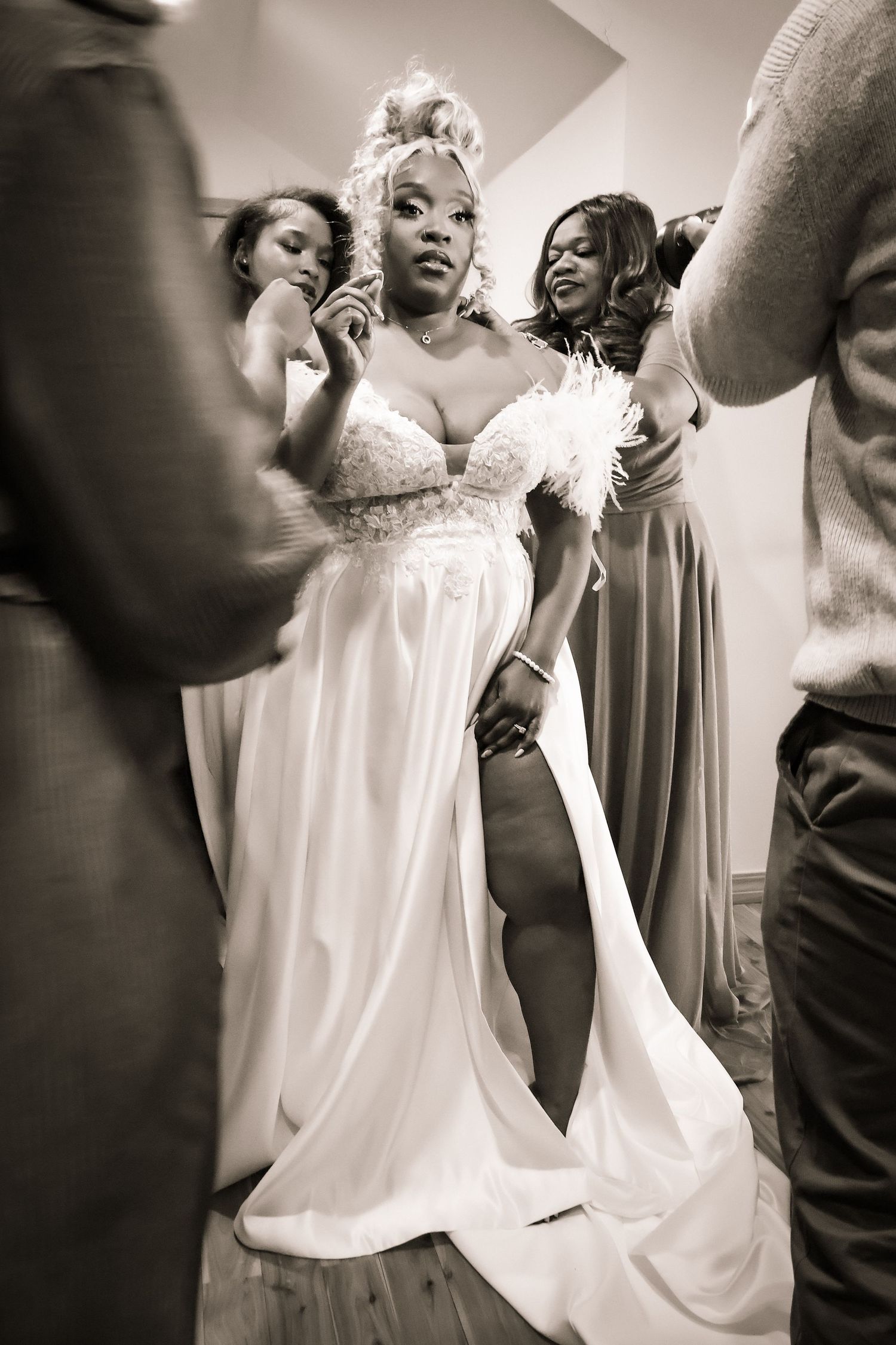 Bride in elegant white gown surrounded by bridesmaids helping her prepare in black and white photo.