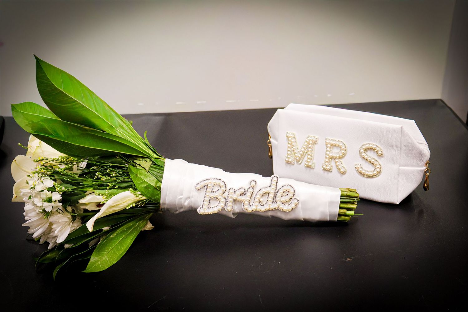 Bridal bouquet of white flowers wrapped with Bride ribbon next to a MRS clutch purse.