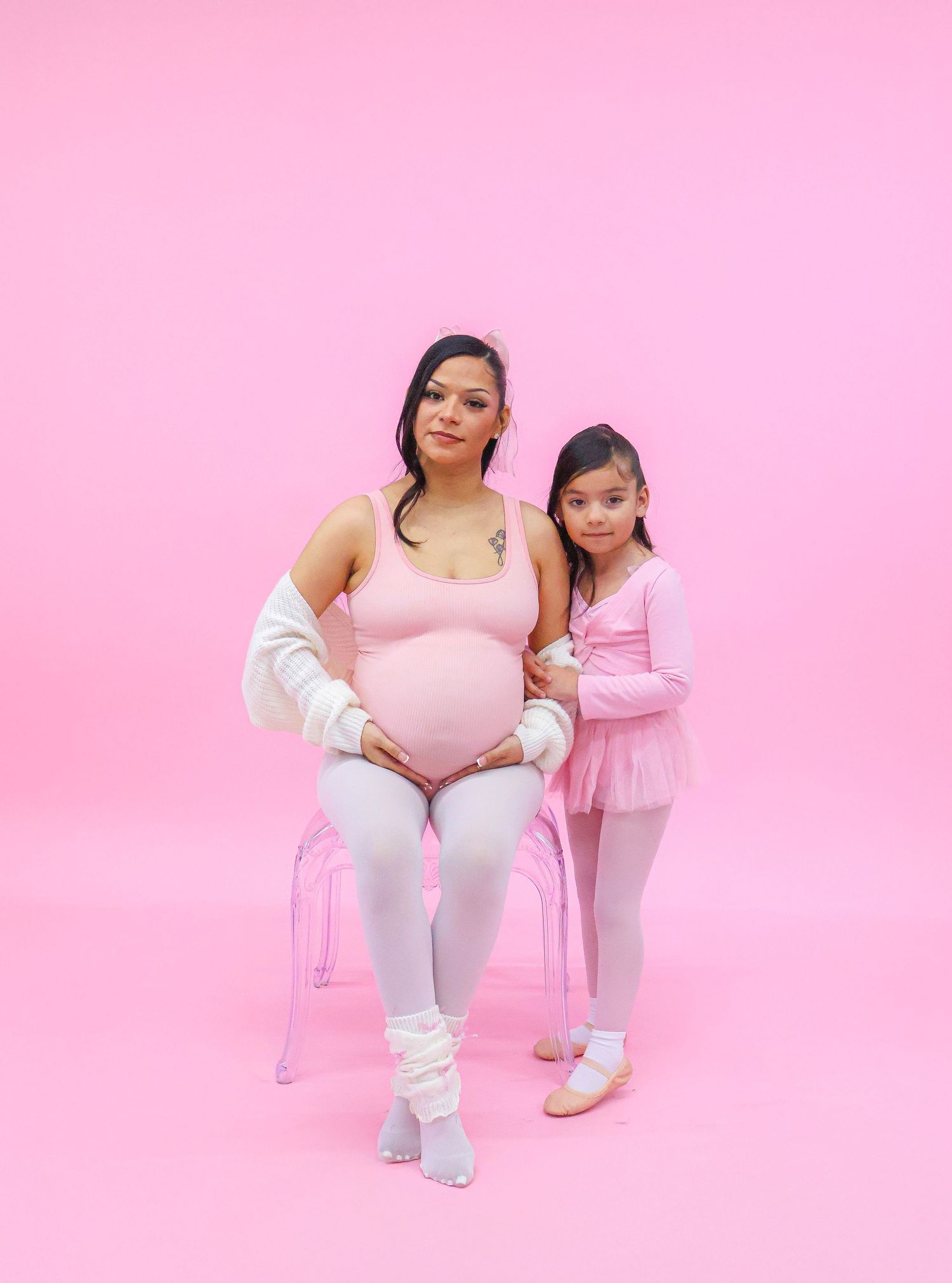 Pregnant woman in pink bodysuit sits beside young girl in ballet attire against a pink background.