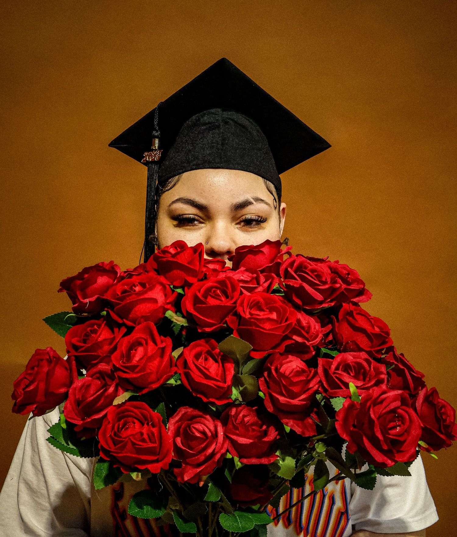 Graduate in black cap peers over large bouquet of red roses on golden background.