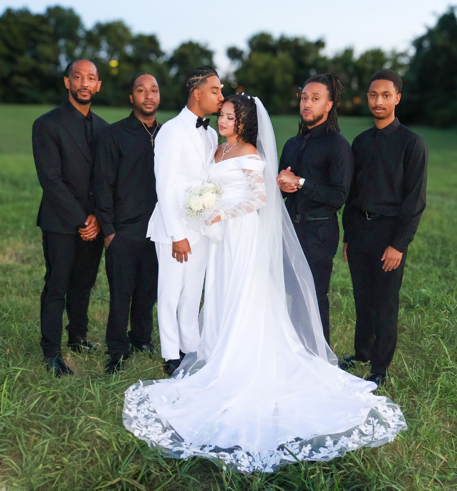 Wedding couple kissing in a field, surrounded by four groomsmen in black attire, bride in white gown with veil.