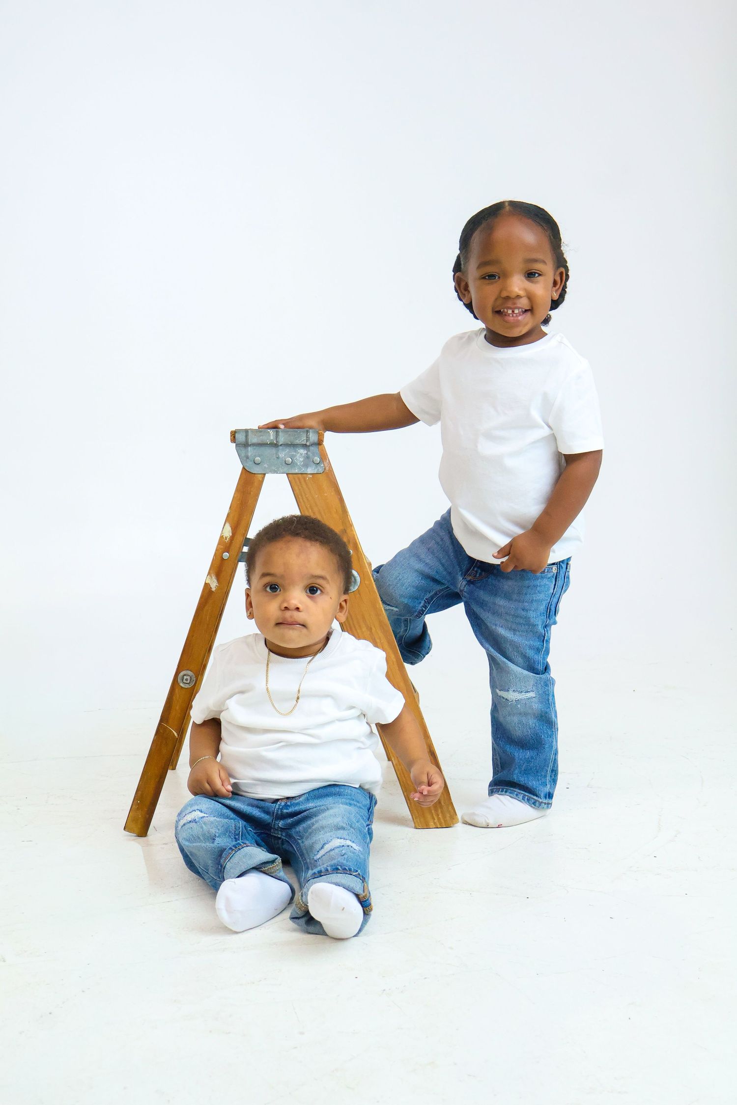 Two young Black boys in matching white tees and jeans pose with a wooden step ladder on a white background.