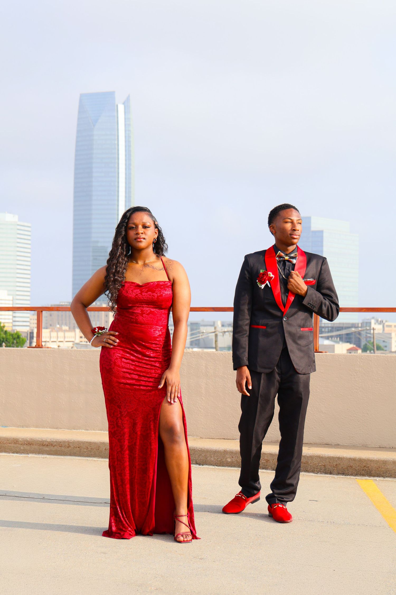 A couple in matching red and black formal wear poses on a rooftop with a city skyline backdrop.