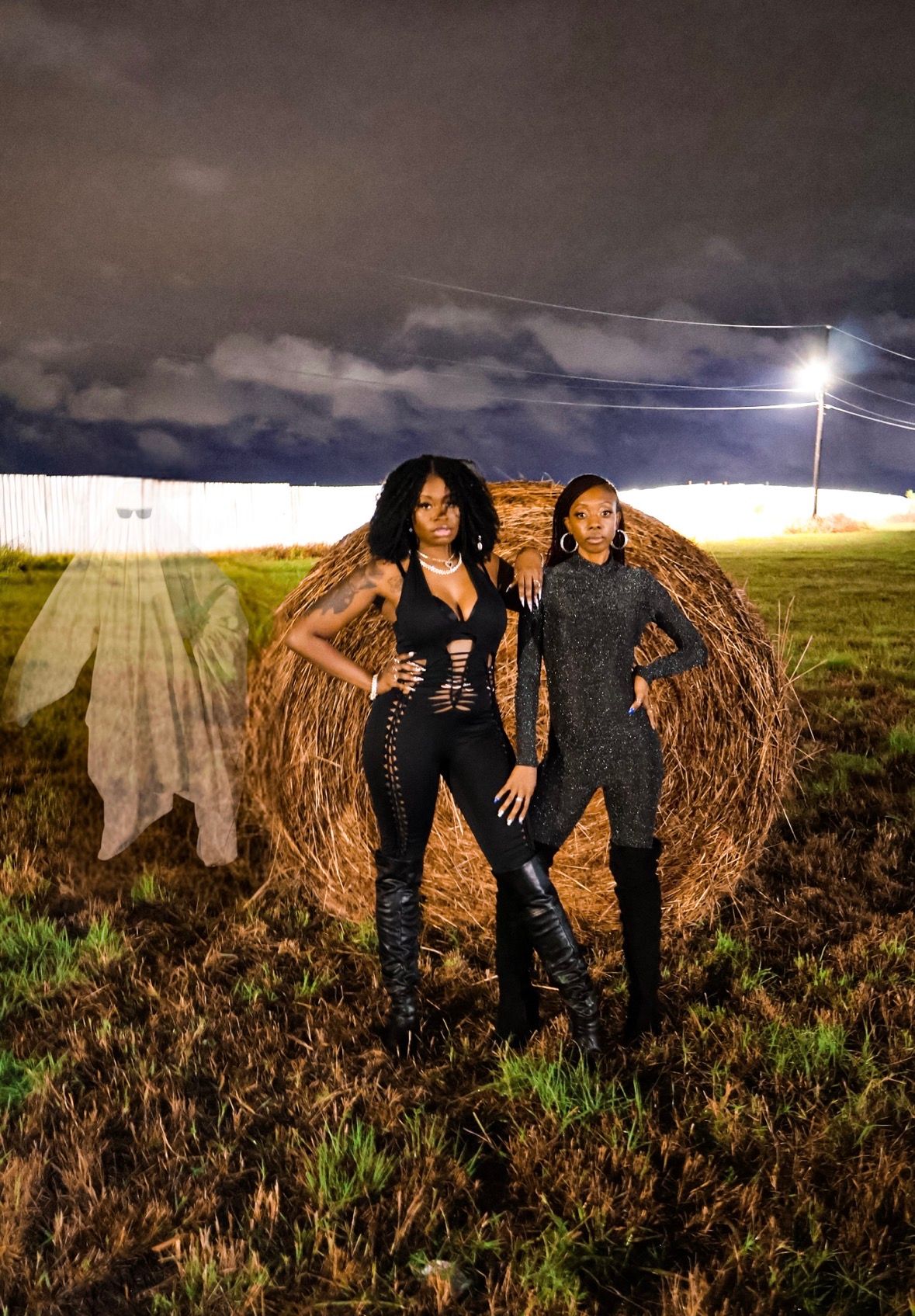 Two women in black outfits stand before a hay bale at night with a ghostly figure nearby.