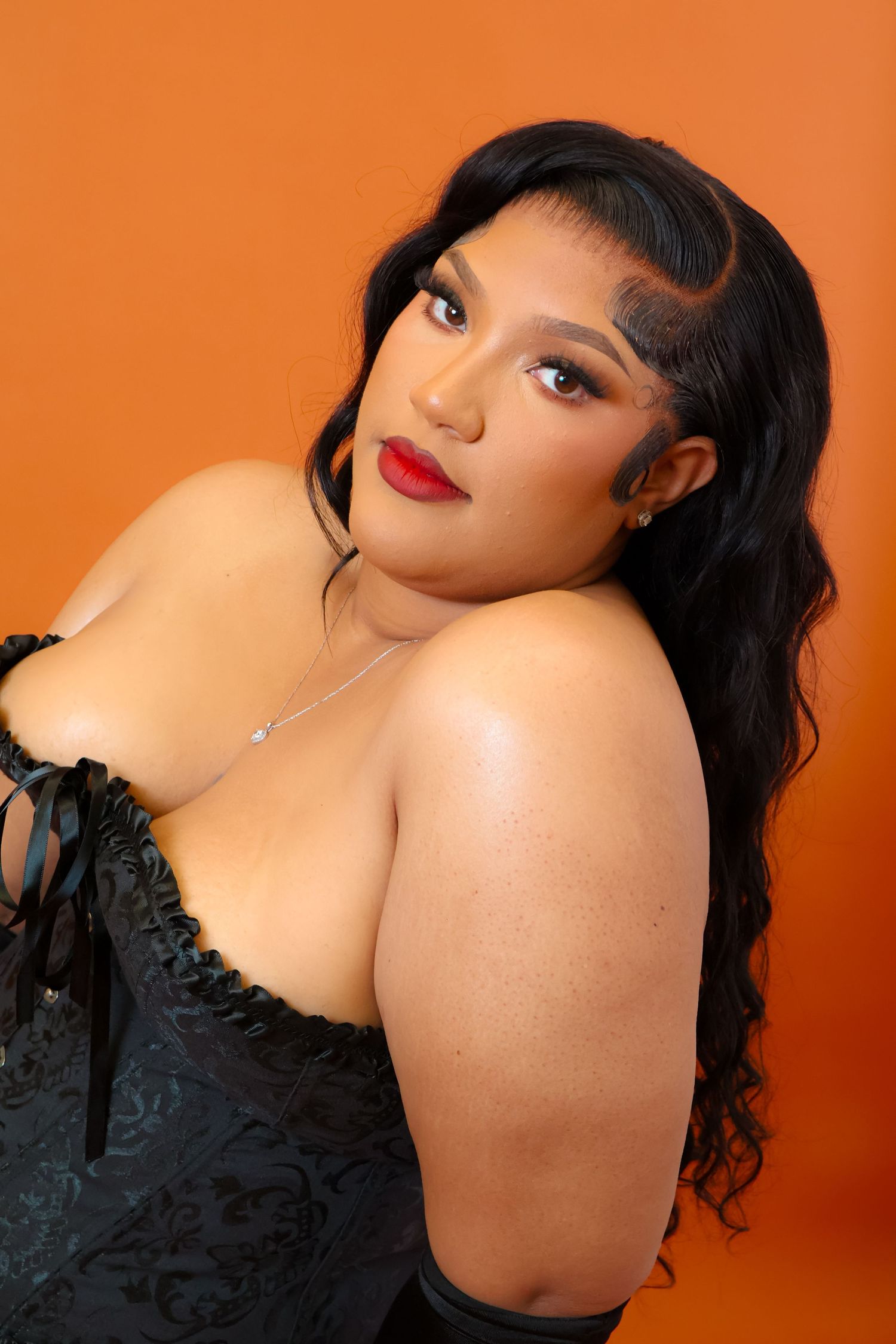 Young woman in black lace corset with red lips and wavy hair posing against vibrant orange background.