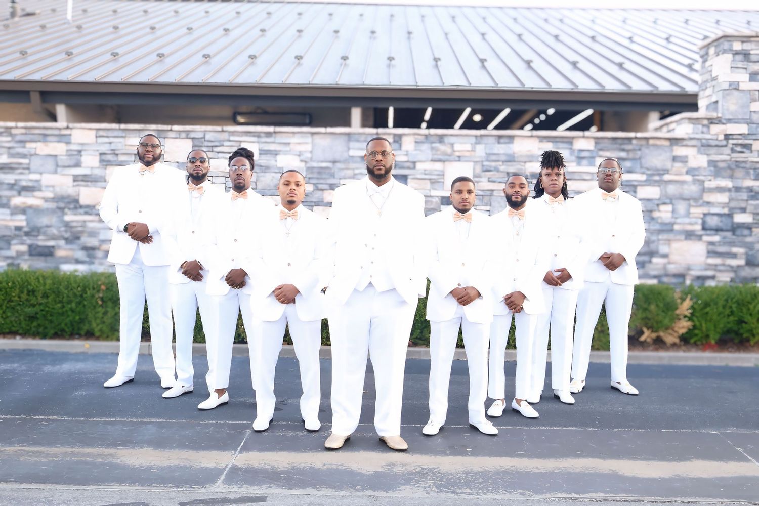 Groom and groomsmen dressed in matching all-white suits pose together outside wedding venue.