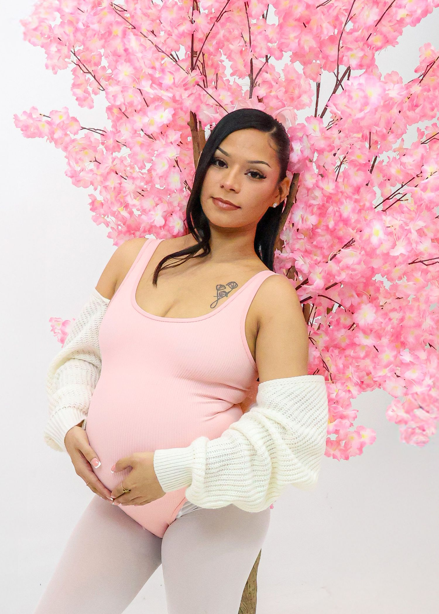 Pregnant woman in pink bodysuit and white cardigan posing before a pink cherry blossom tree backdrop.
