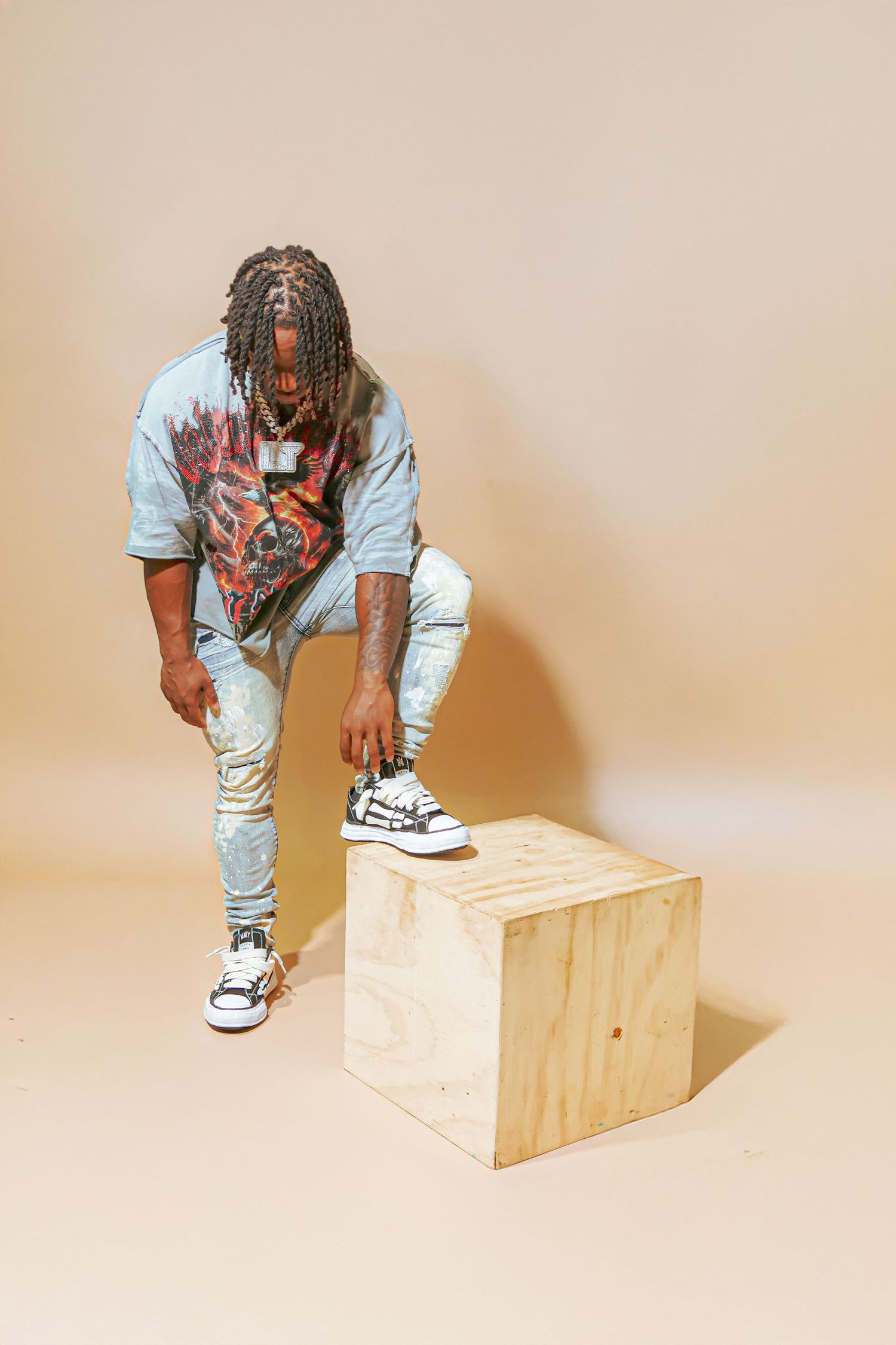 Young man with dreadlocks in light blue jeans and graphic tee, posing with foot on wooden box.