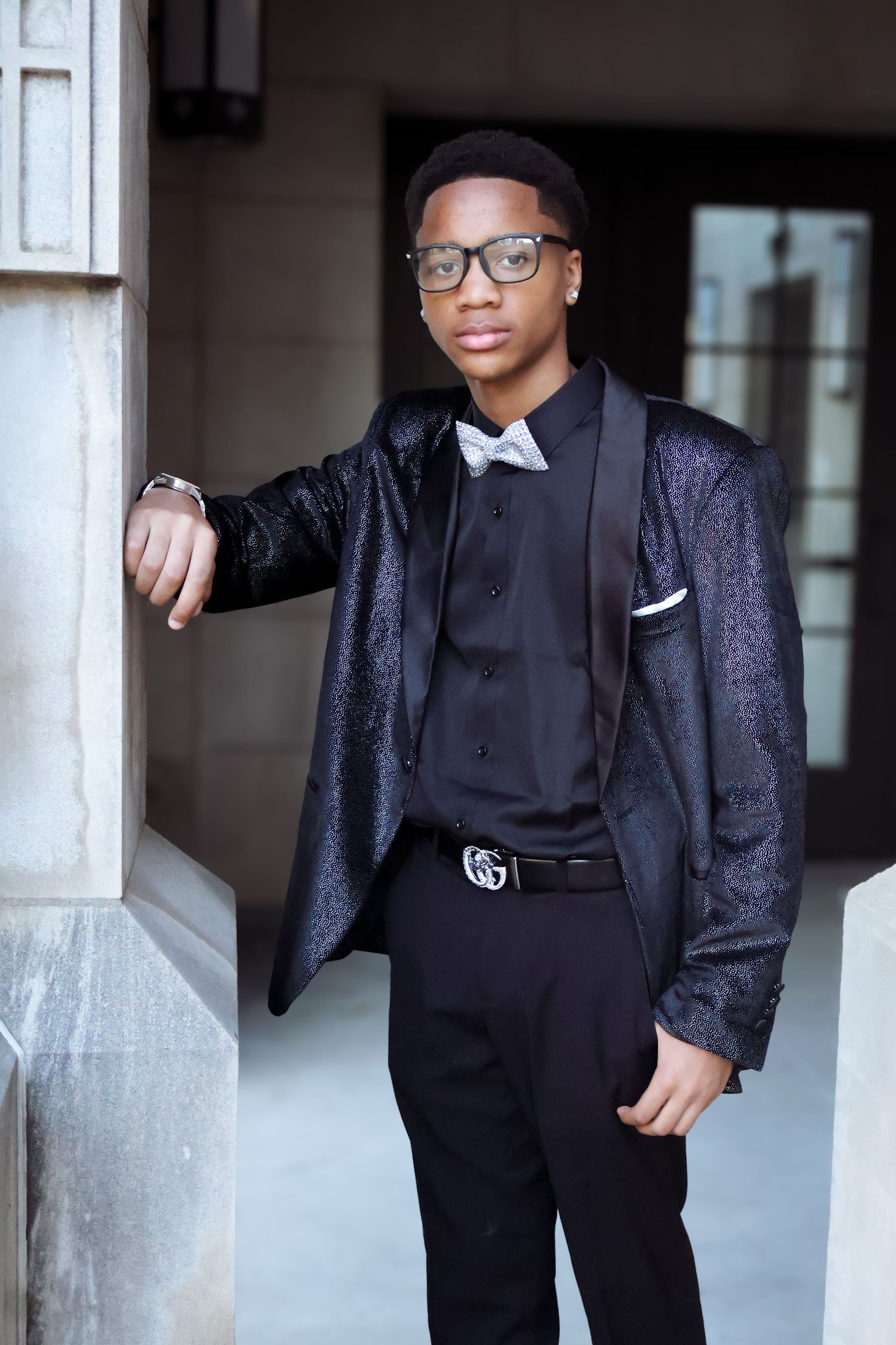 Stylish young man in black sequin blazer and bow tie leaning against stone column.