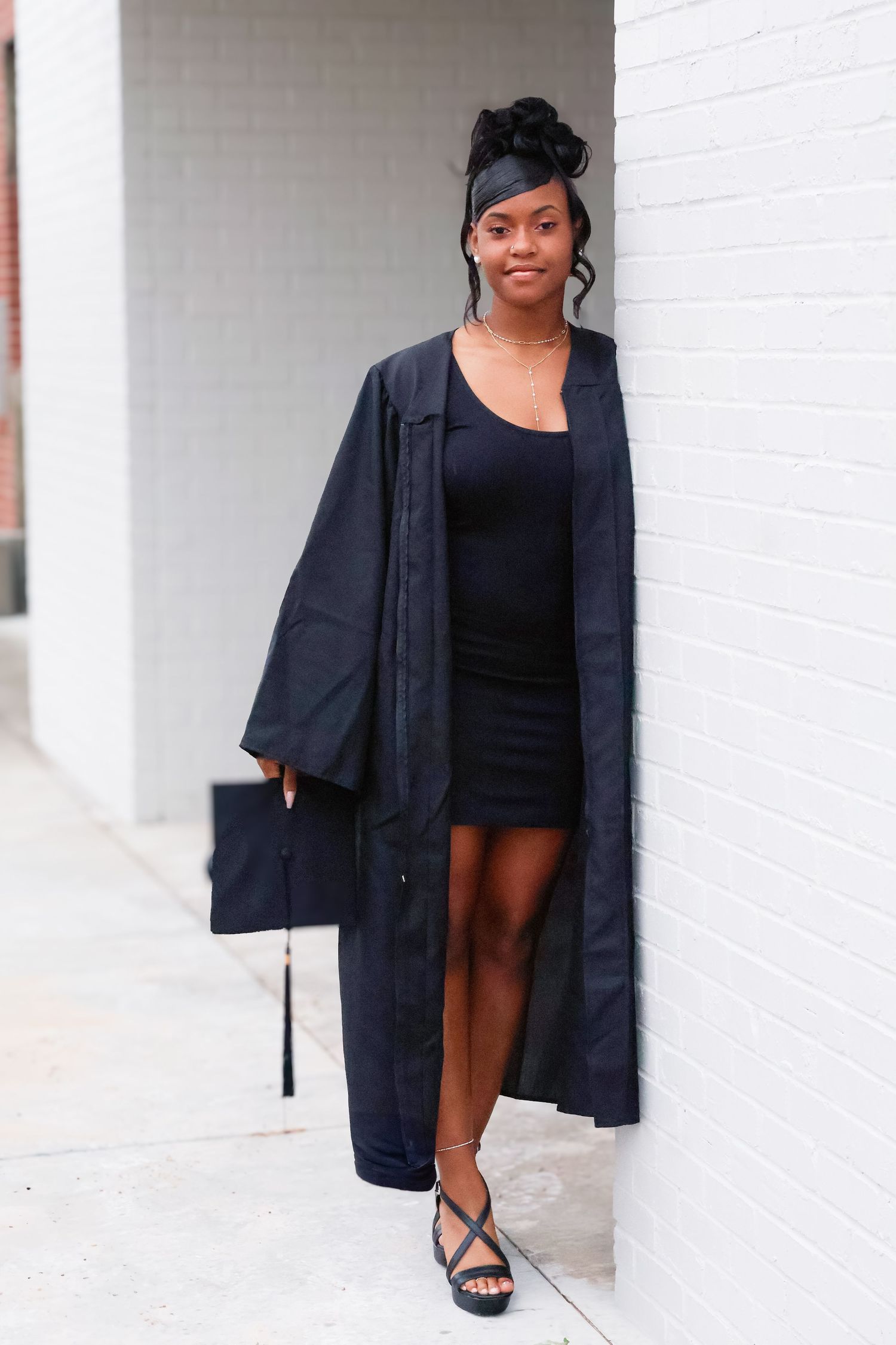 Young woman in black graduation cap and gown leaning against white brick wall outdoors.