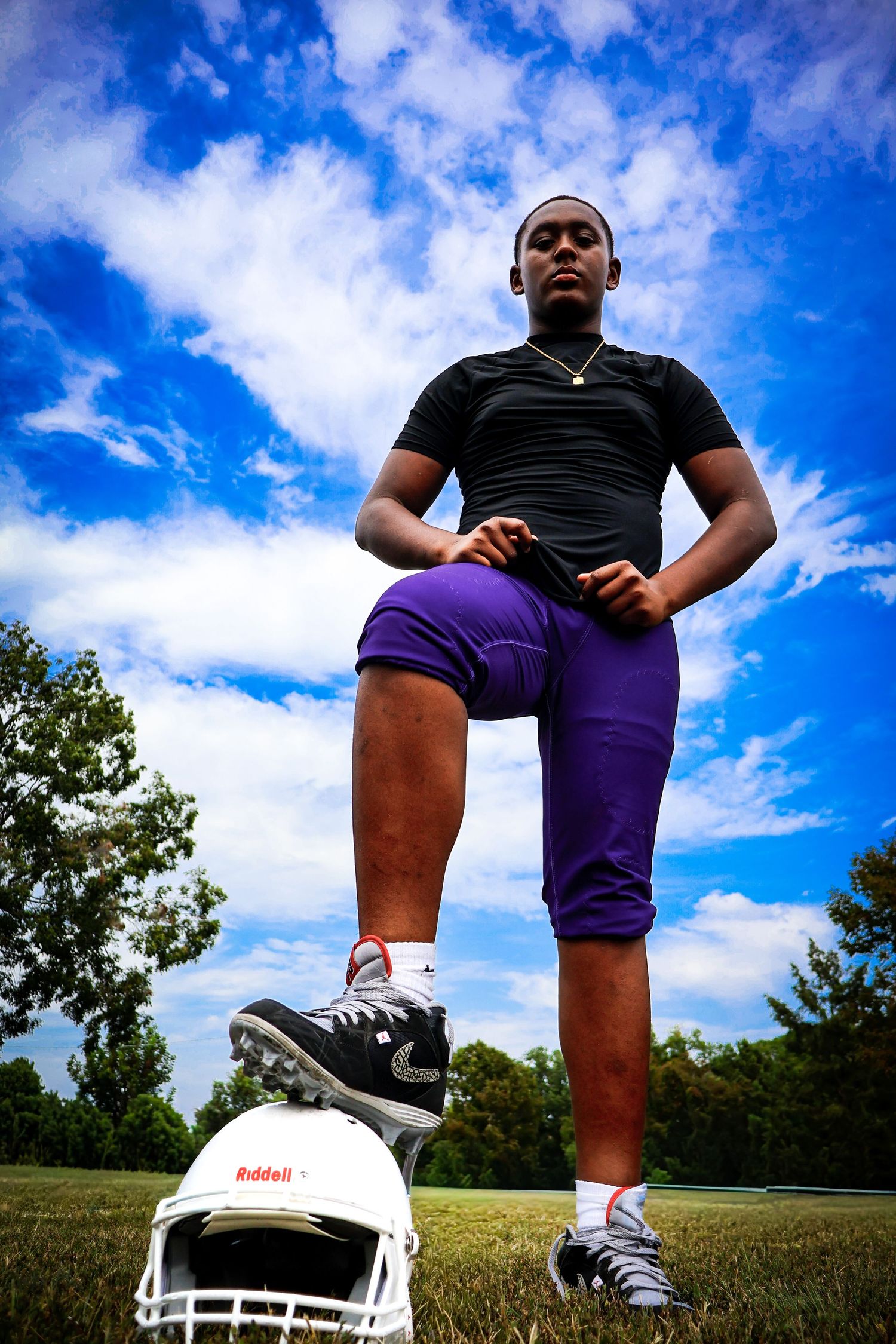 A young football player in purple and black uniform poses confidently with foot on helmet outdoors.