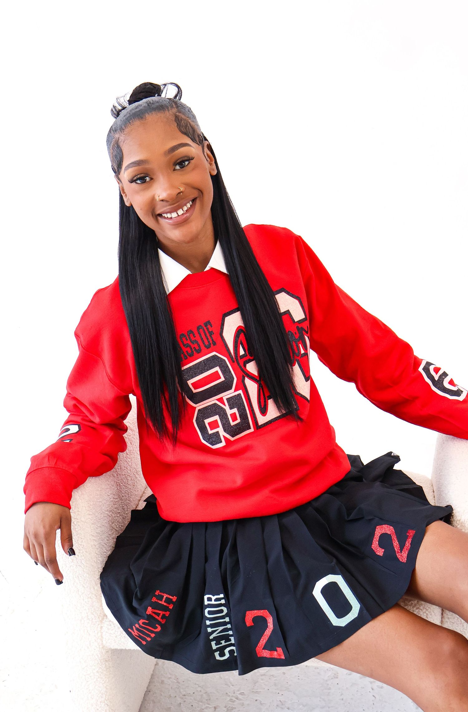 Smiling young woman in red Senior 2023 sweatshirt and navy pleated skirt sitting on white chair.