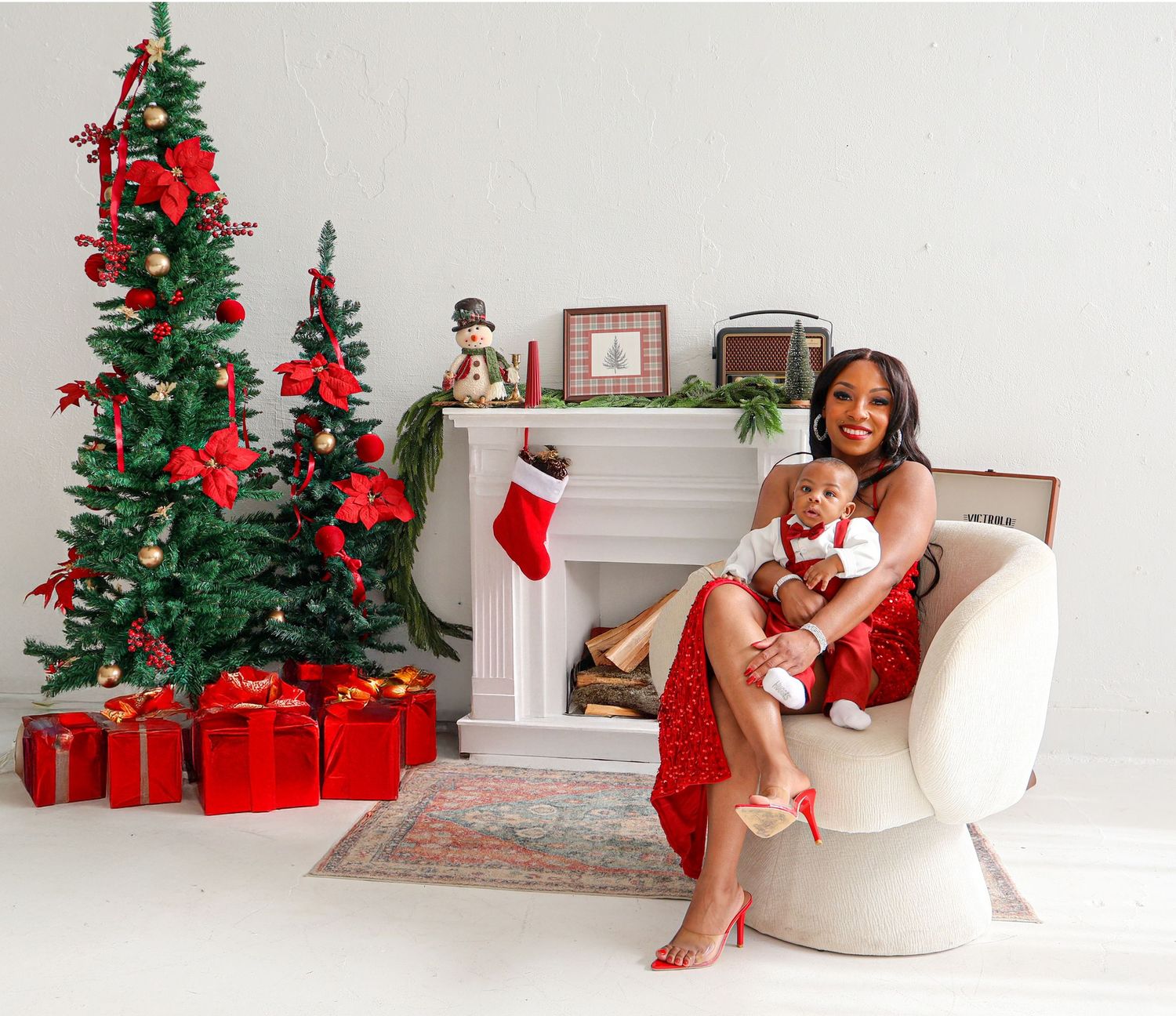 Woman in red dress holds baby on chair beside decorated Christmas tree with gifts and white fireplace.