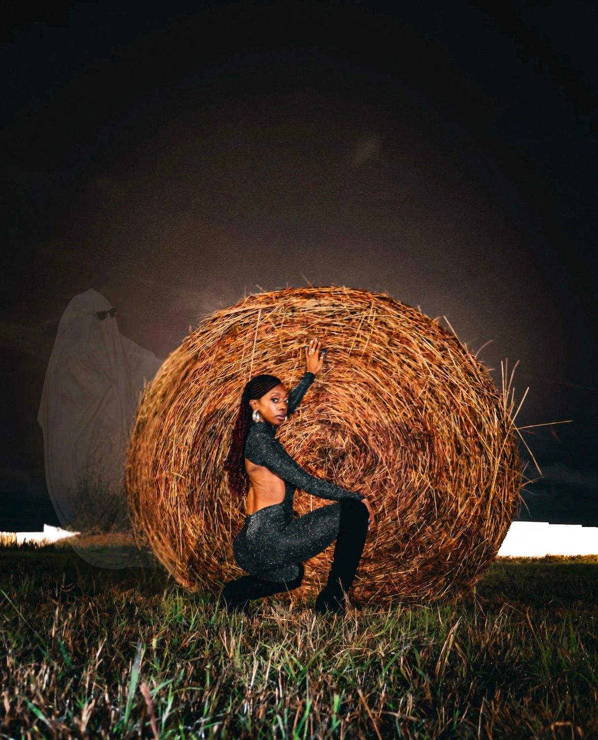 A woman in a black outfit poses beside a hay bale at night with a ghost figure behind her.