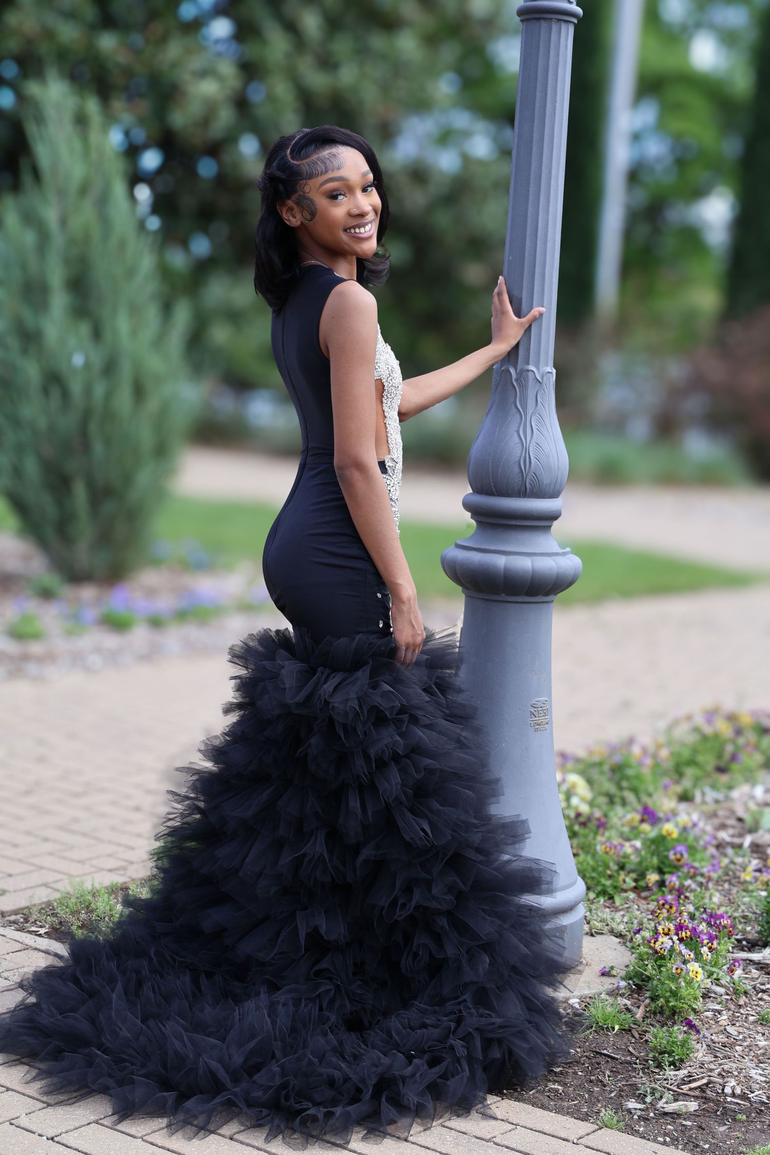 Young woman in elegant black mermaid prom gown with tulle train poses by decorative lamp post outdoors.