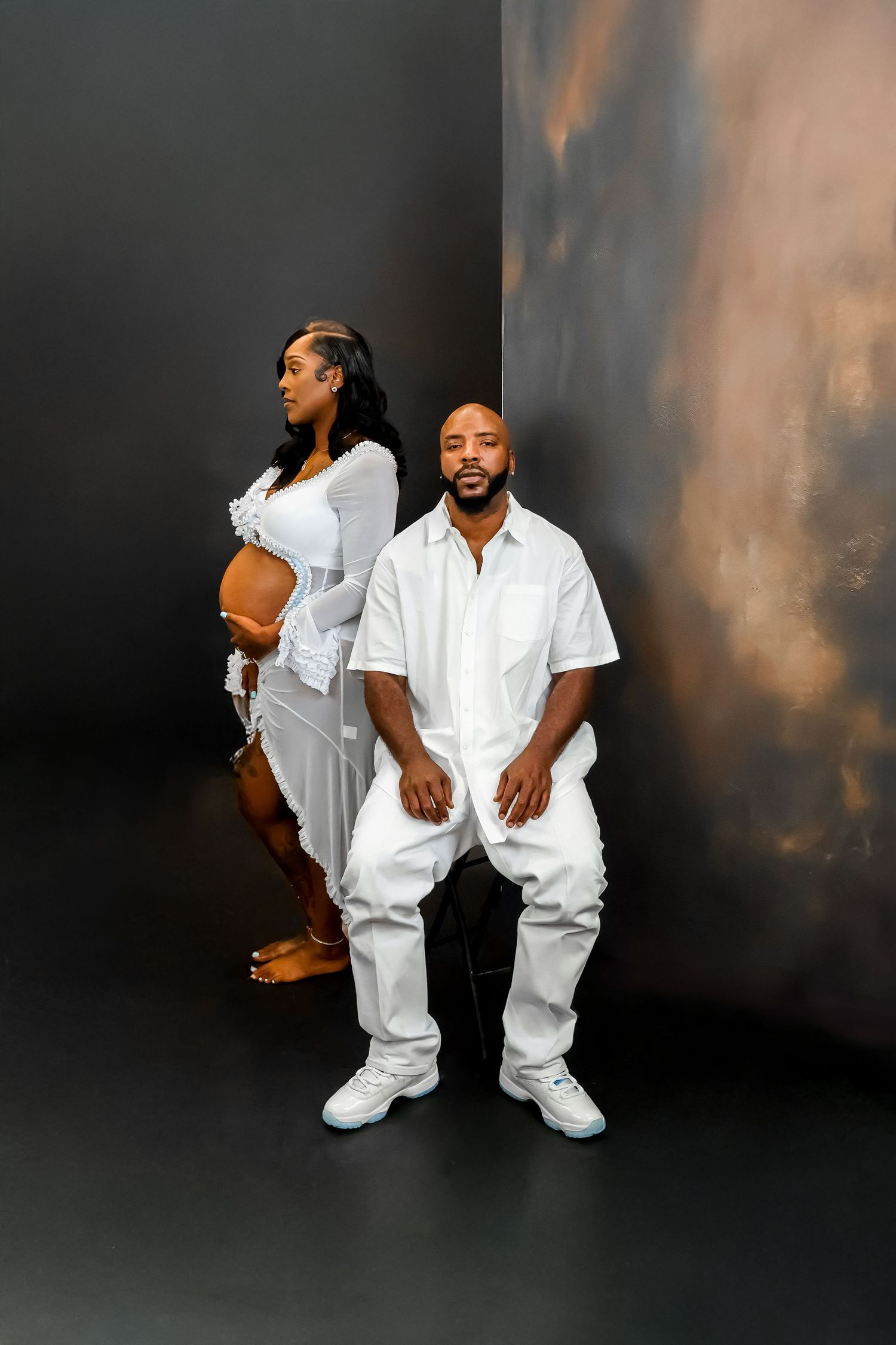 A couple in matching white outfits poses for a maternity photo shoot against a dark studio backdrop.