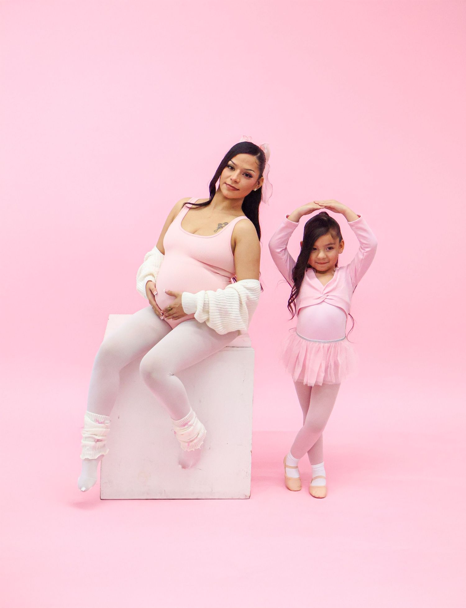 Pregnant woman in pink outfit sits on white cube beside young girl in ballet attire against pink background.