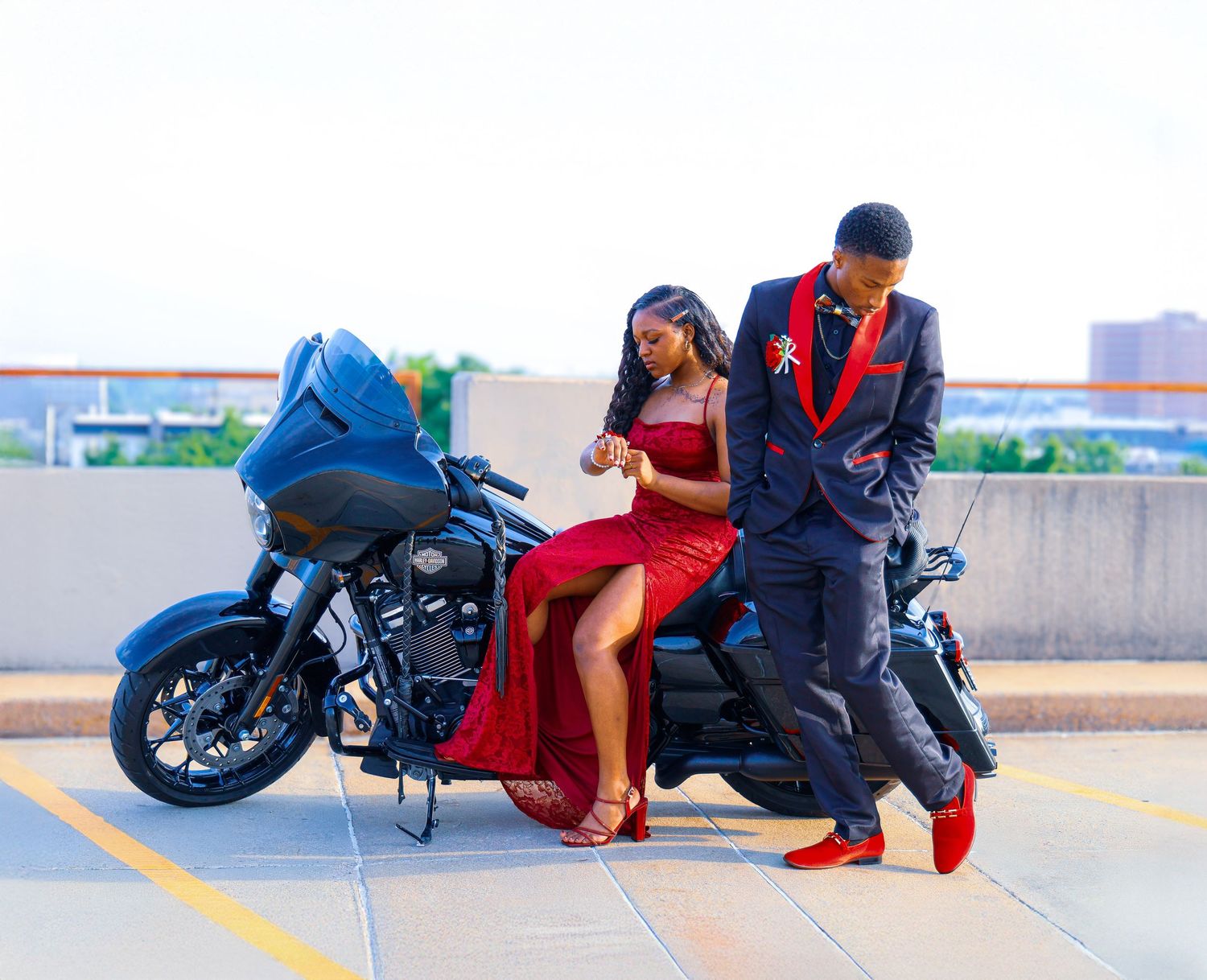 Couple in formal prom attire posing beside a Harley-Davidson motorcycle on a rooftop parking lot.