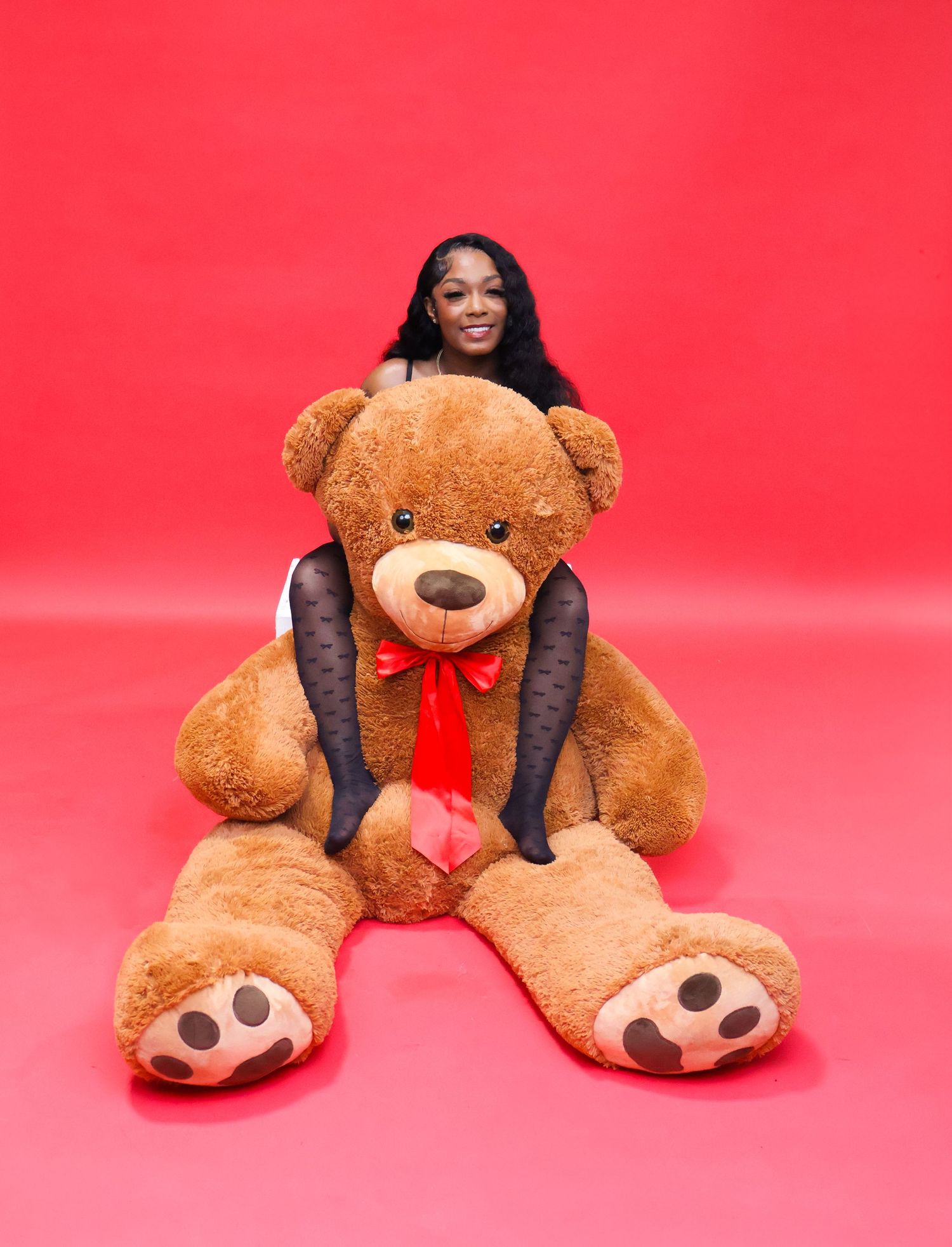 Woman smiling behind a giant brown teddy bear with red bow on a vibrant red background.