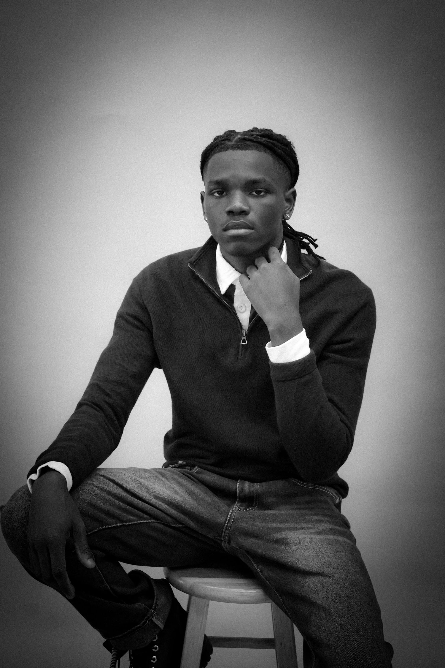 Young man with braids sits thoughtfully on stool in black and white portrait photo.
