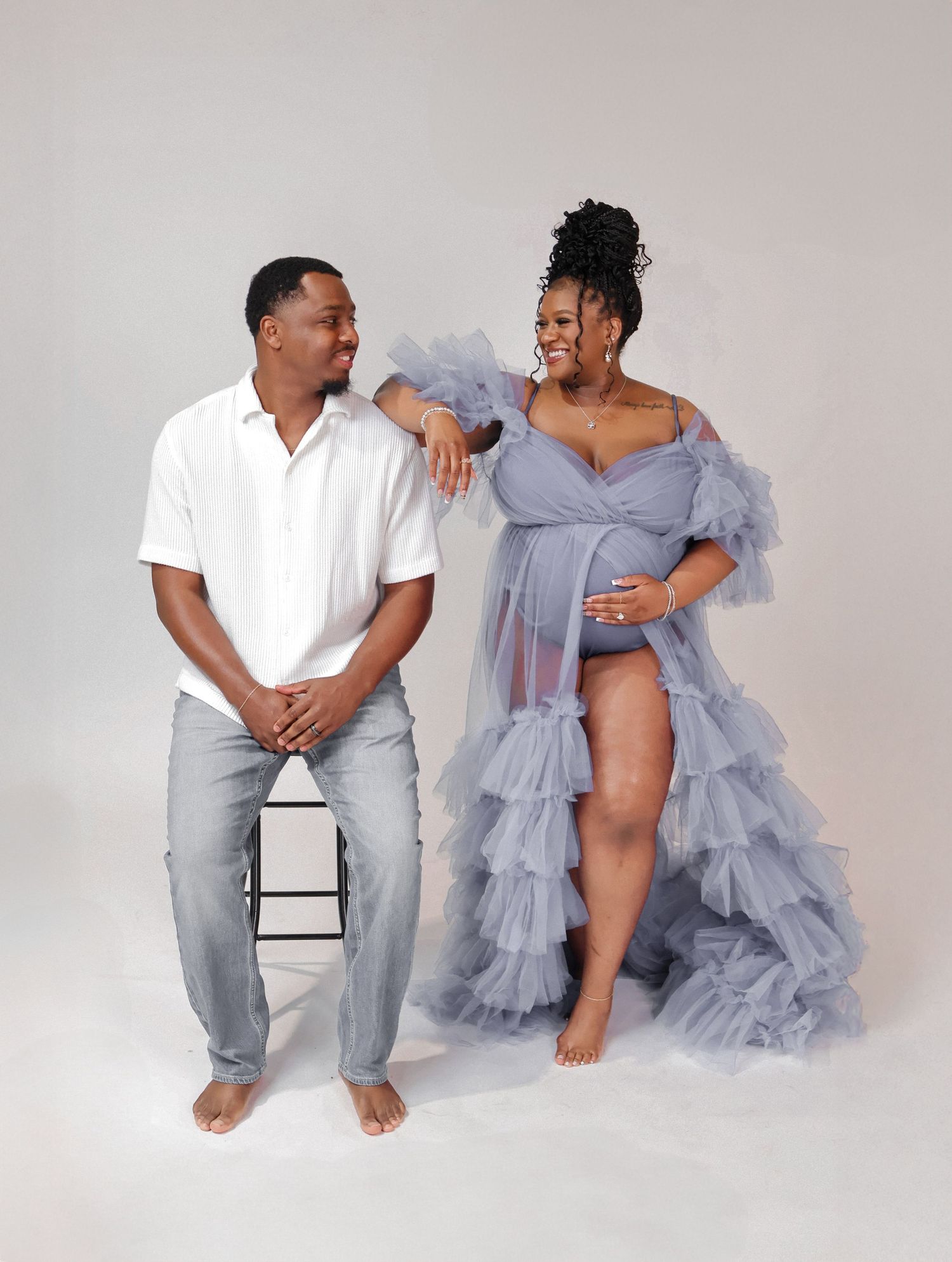 Couple poses for maternity photo shoot, woman in blue tulle gown cradles baby bump beside man in white shirt and jeans.
