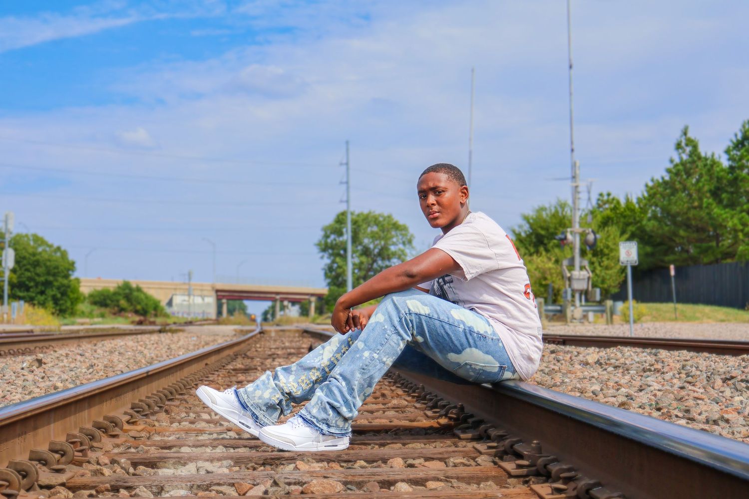 Young Black man in a white t-shirt and jeans sitting on railroad tracks under a blue sky.
