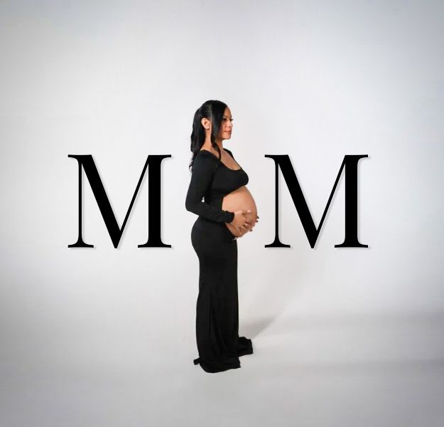 Pregnant woman in black gown poses sideways between large MOM letters on white background.