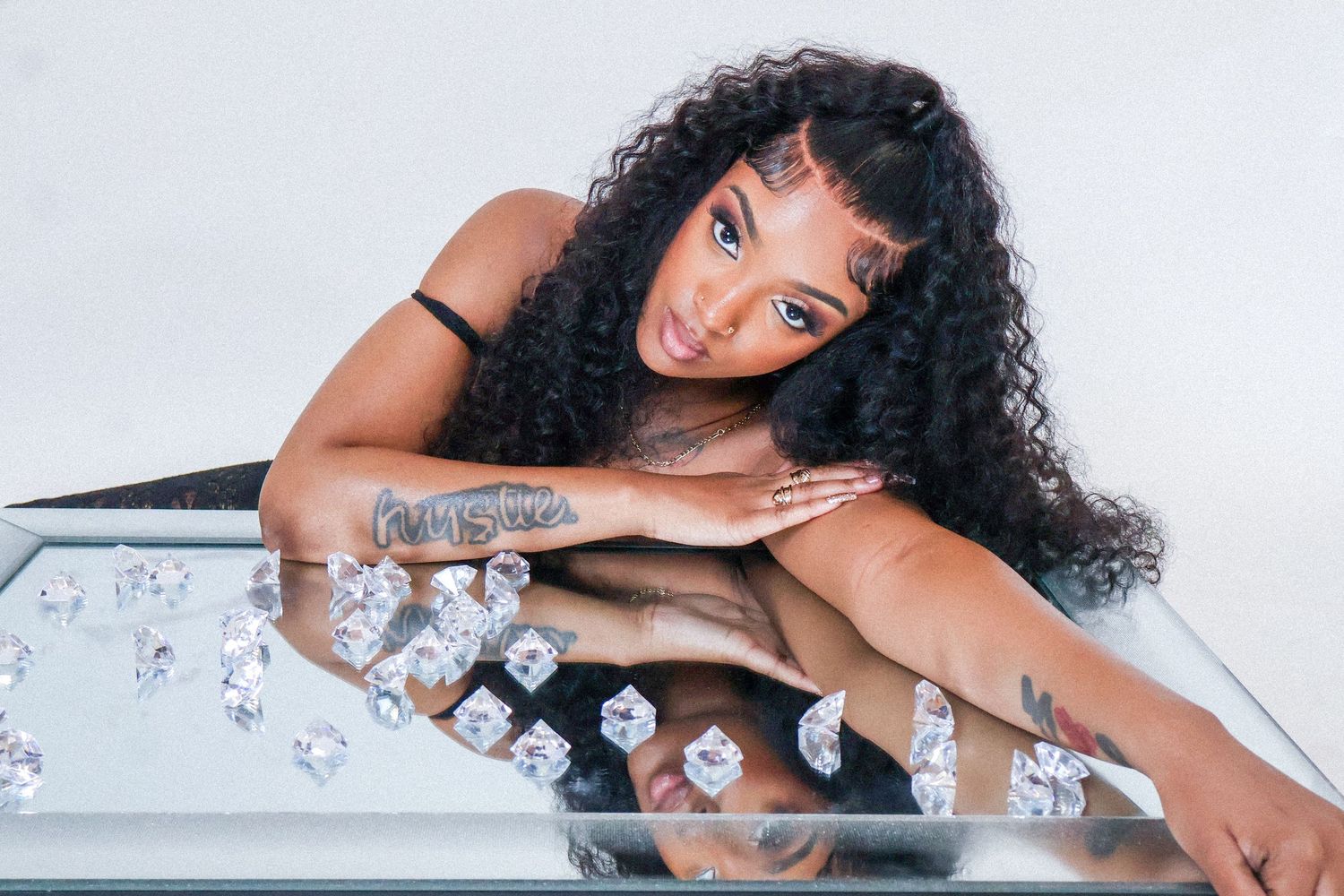A woman with long curly hair leans on a mirrored surface surrounded by scattered diamonds in a stylish photo shoot.