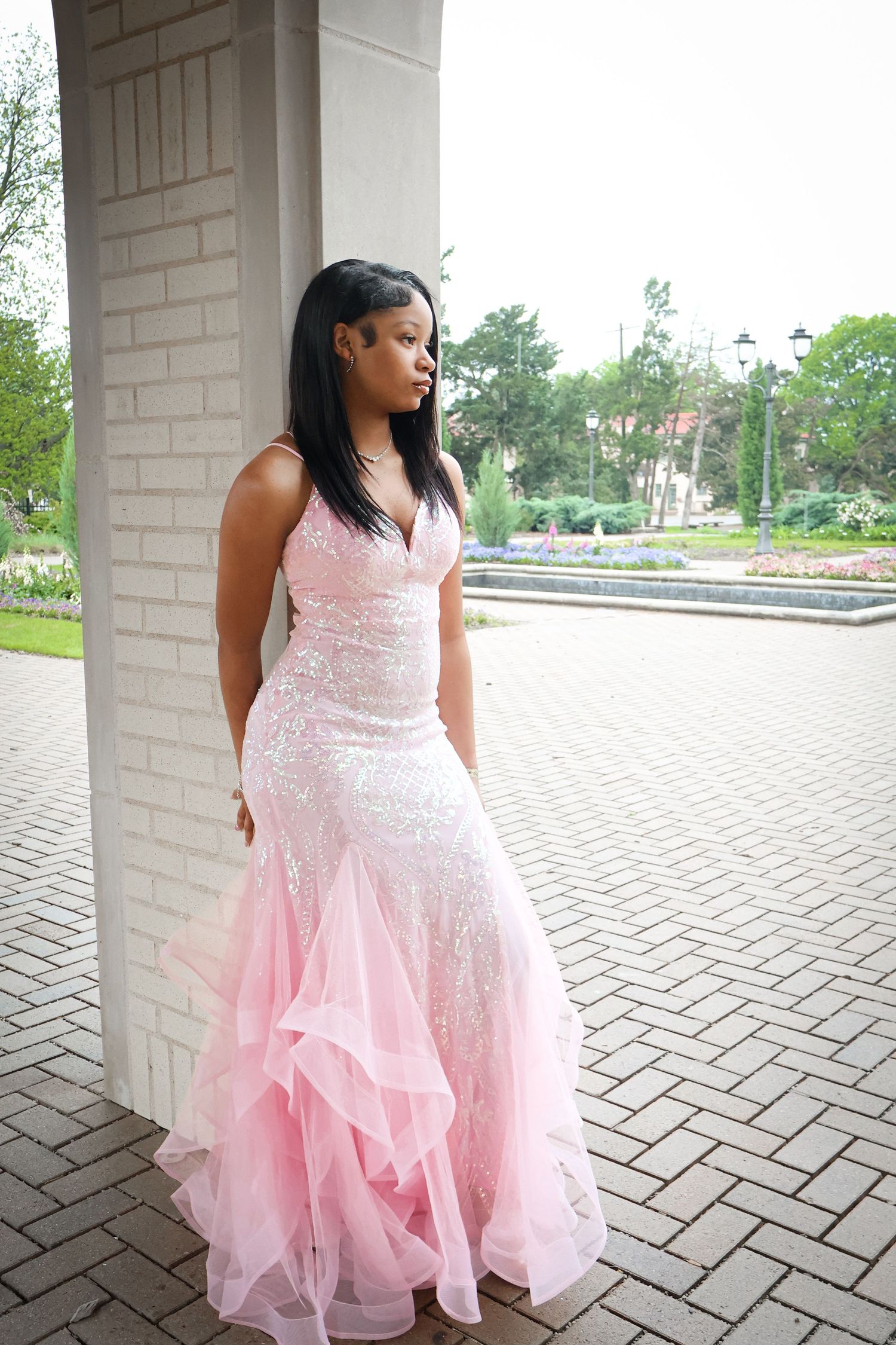 Young woman in pink sequin mermaid prom dress posing beside white column in garden.