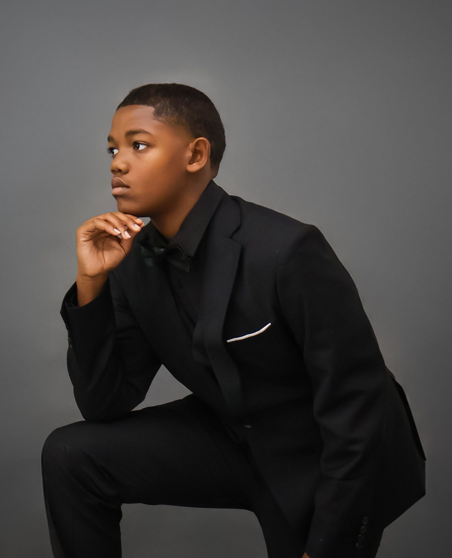 Young boy in black suit sitting thoughtfully with hand on chin.