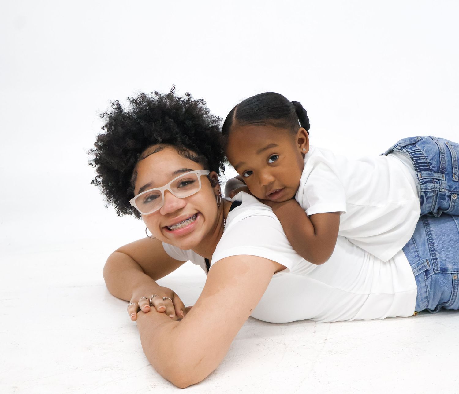 A smiling woman with glasses lies on white floor while a young child in white shirt and jeans leans on her back.