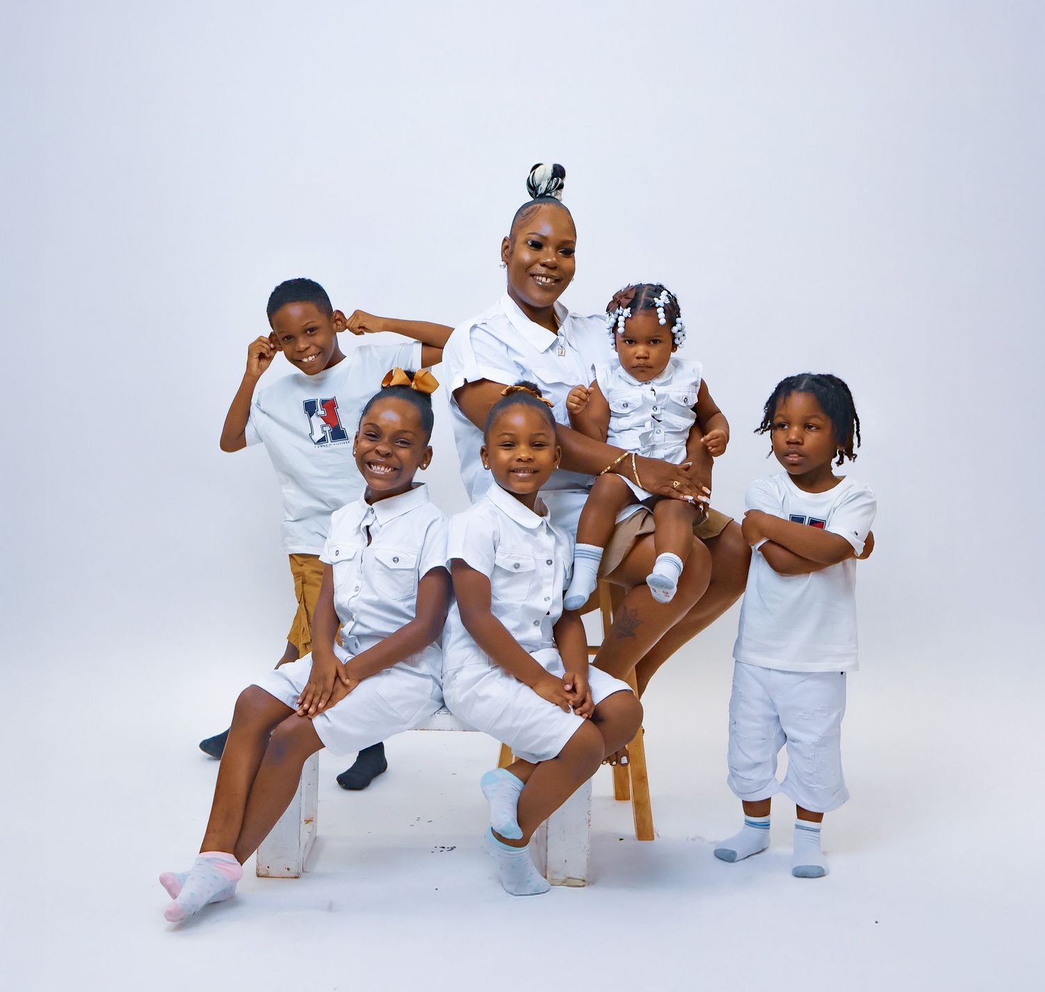 A woman sits on a chair surrounded by five young children, all dressed in matching white outfits.