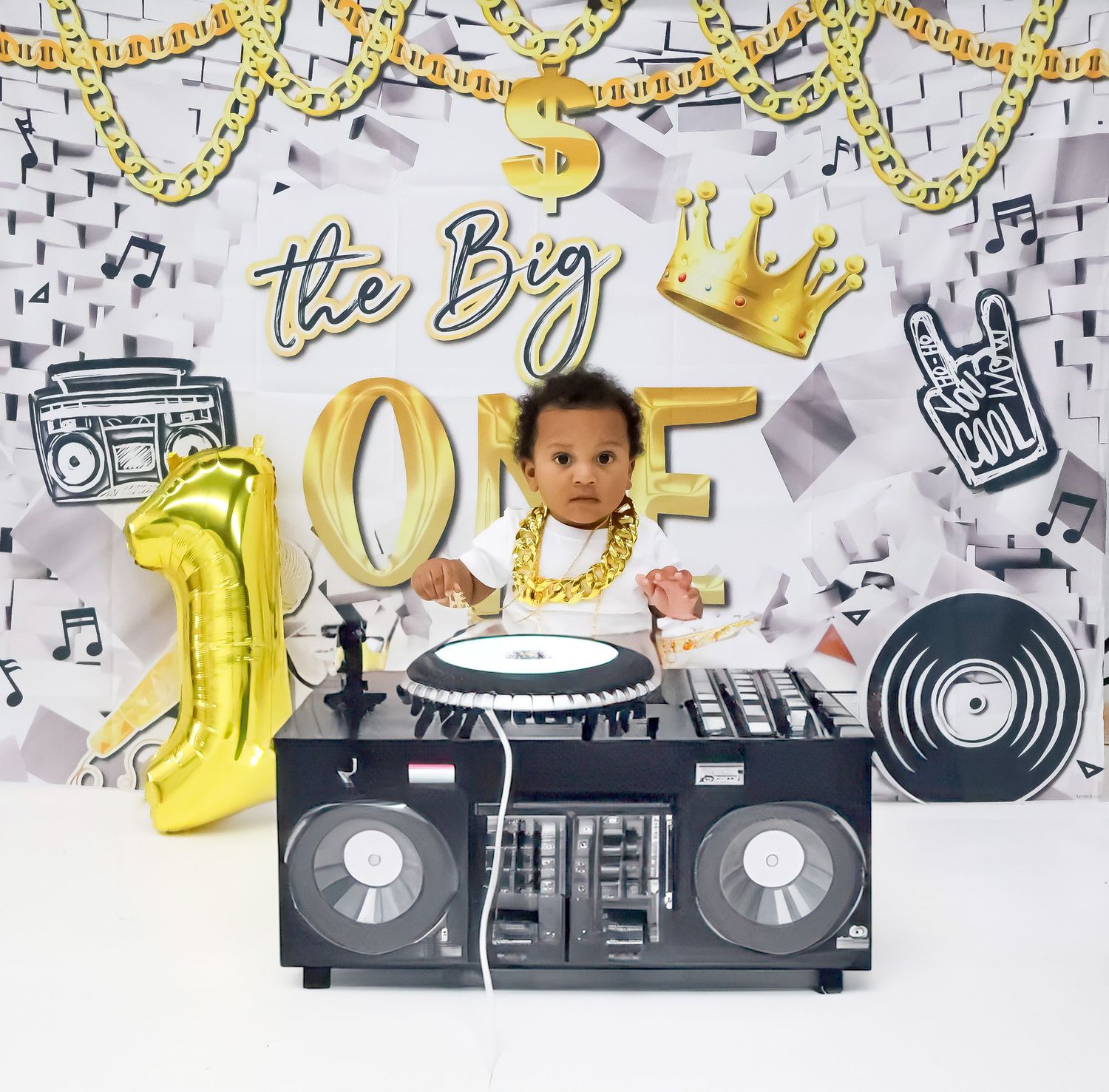 Baby at hip-hop themed first birthday party with DJ setup, gold decorations, and boombox backdrop.