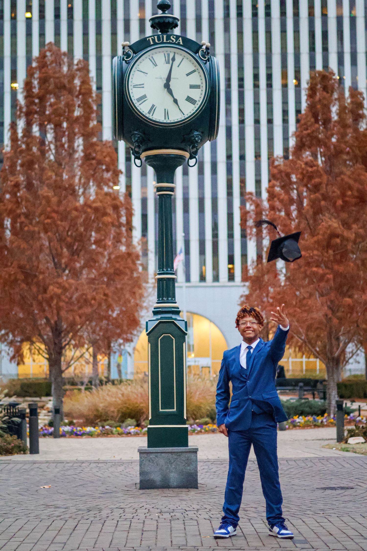 Young Black male graduate in blue suit tosses cap near Tulsa street clock in autumn plaza.