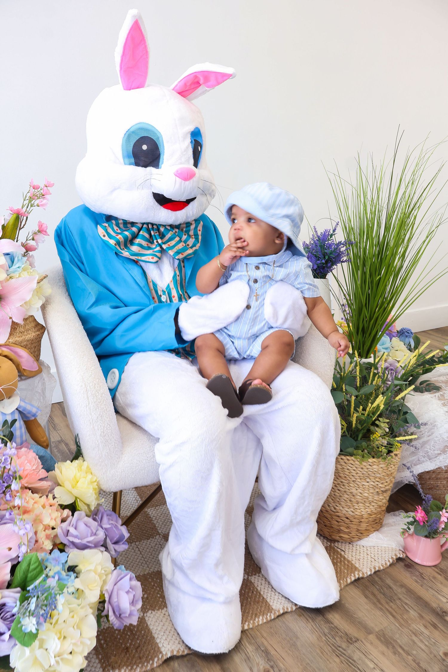Baby in blue outfit sitting on Easter Bunny mascot's lap surrounded by colorful spring flowers.