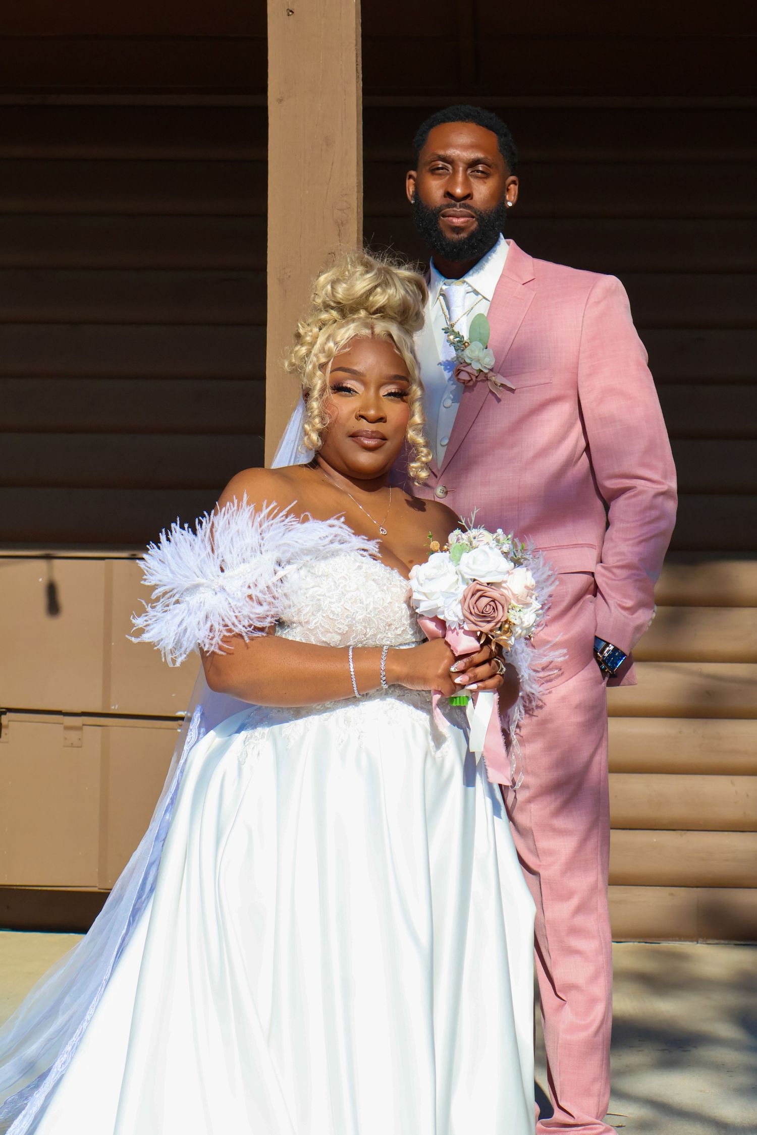 Bride in white feather gown and groom in pink suit pose together at outdoor wedding ceremony.