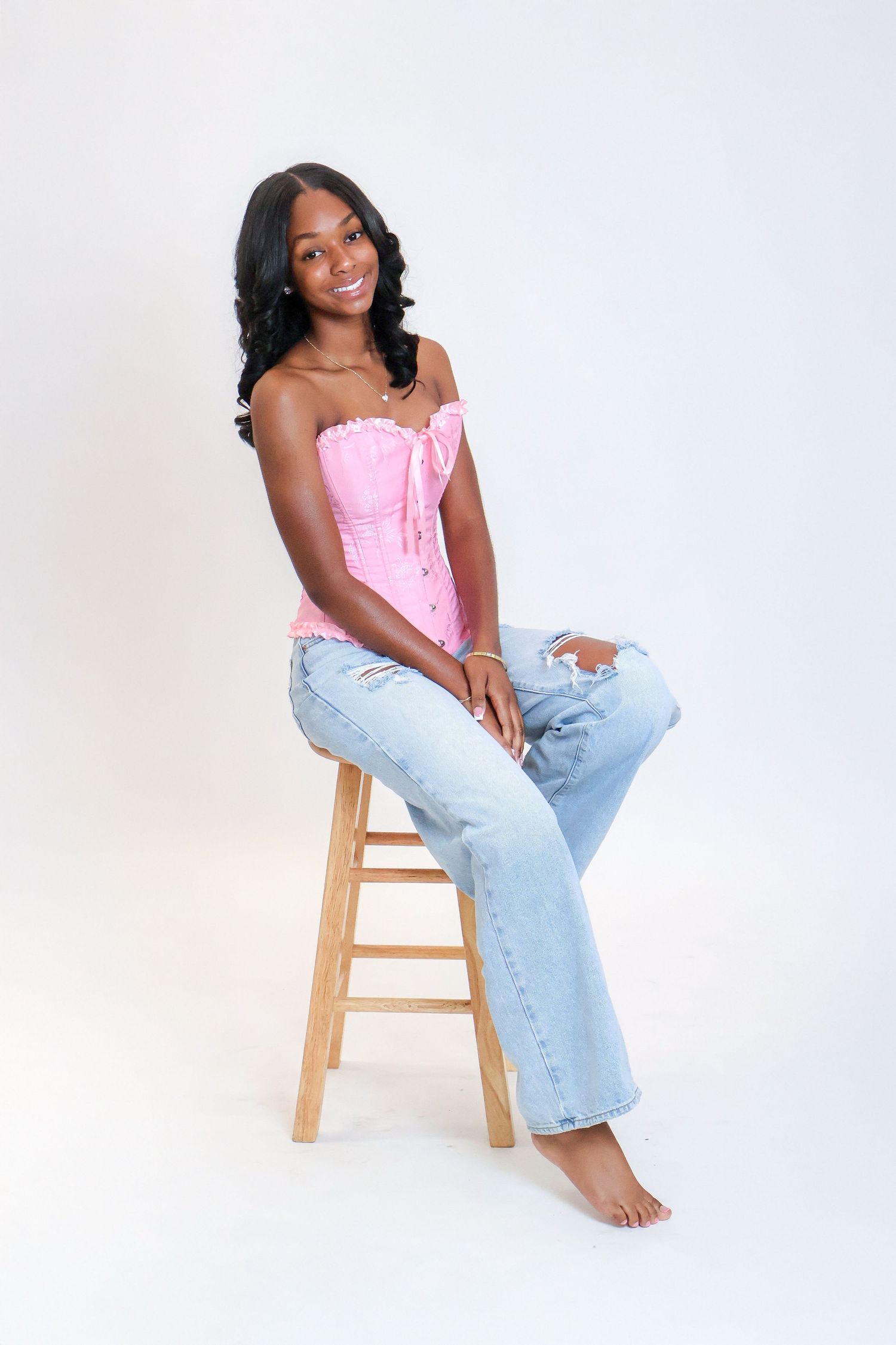 A smiling young woman in a pink corset top and ripped jeans sits barefoot on a wooden stool.