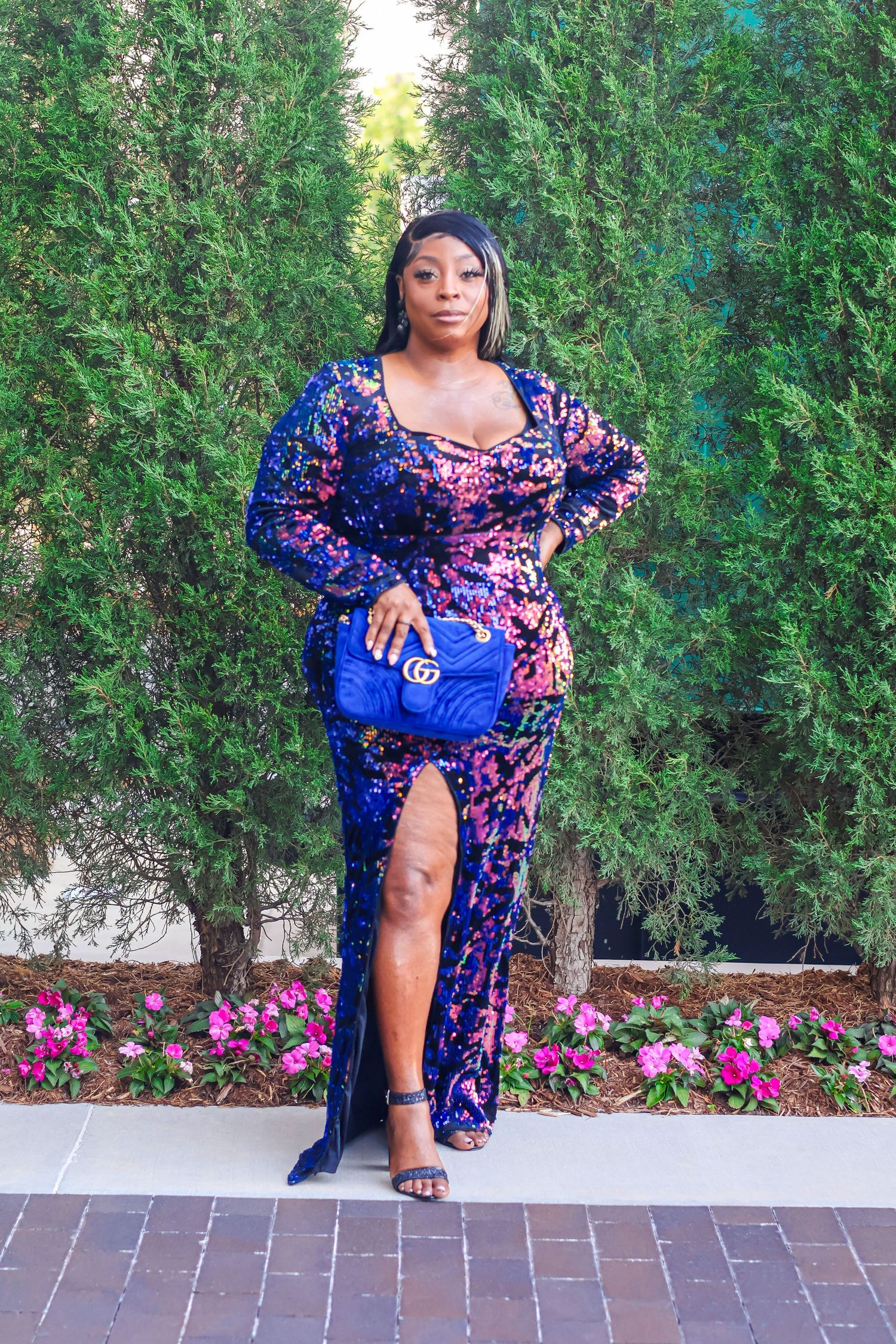 A woman in a blue floral sequined gown carries a Gucci bag while posing among tall green hedges.
