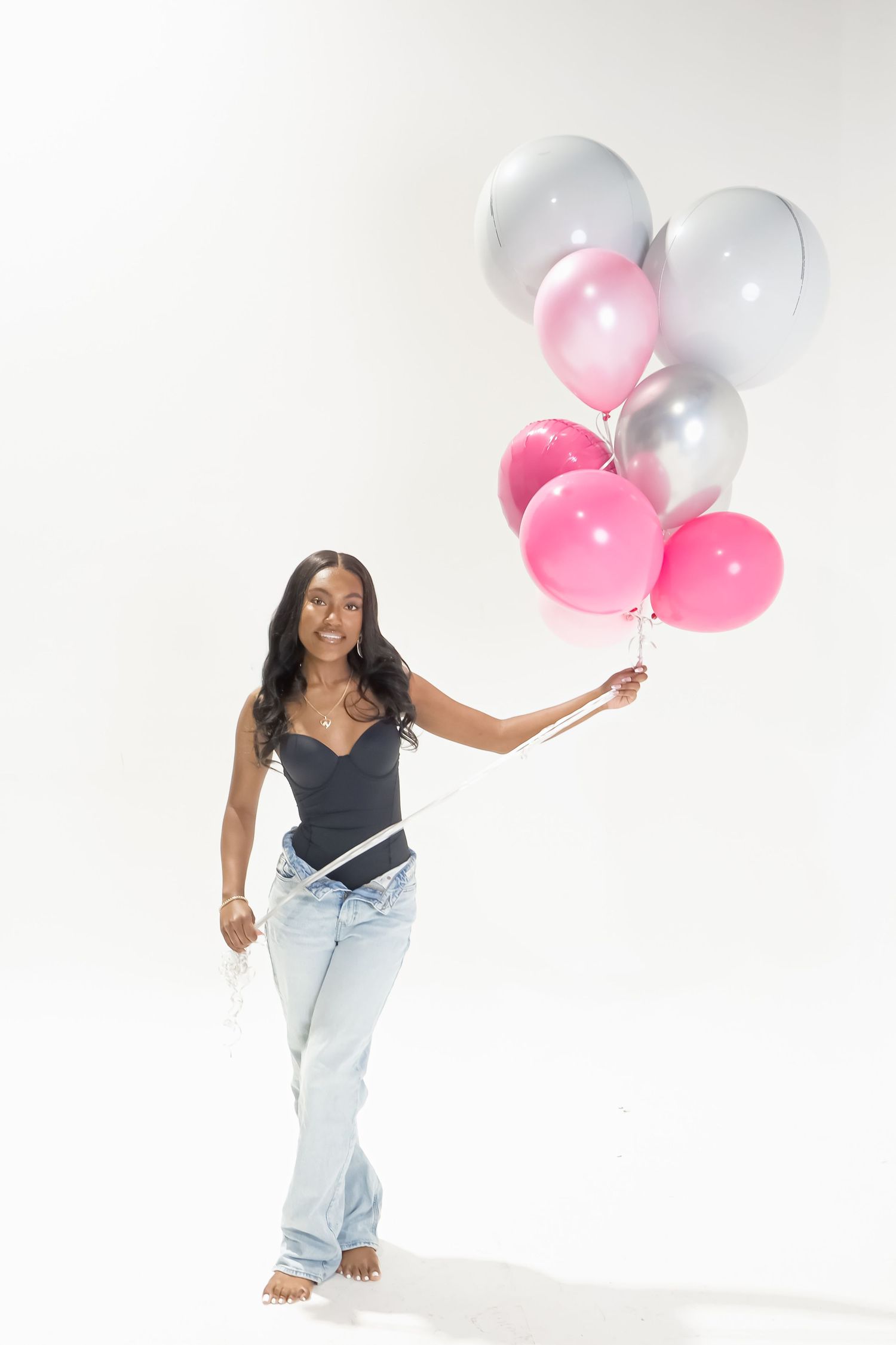 Cheerful young woman in black bodysuit and jeans holding pink and silver balloons on white background.