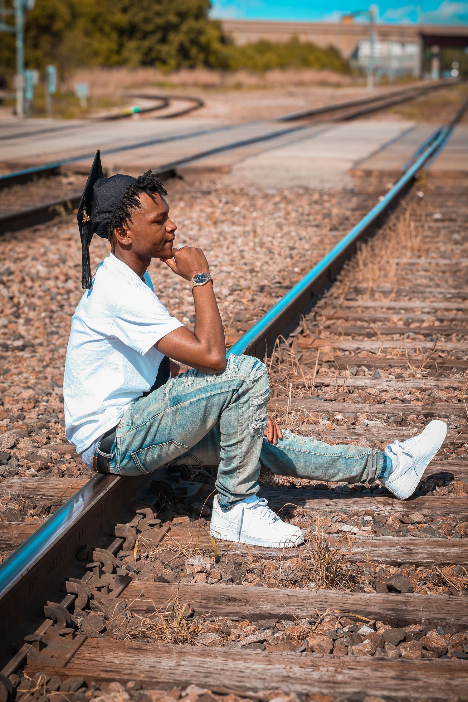 Young man in white tee and jeans sitting on railroad tracks in sunny outdoor setting.