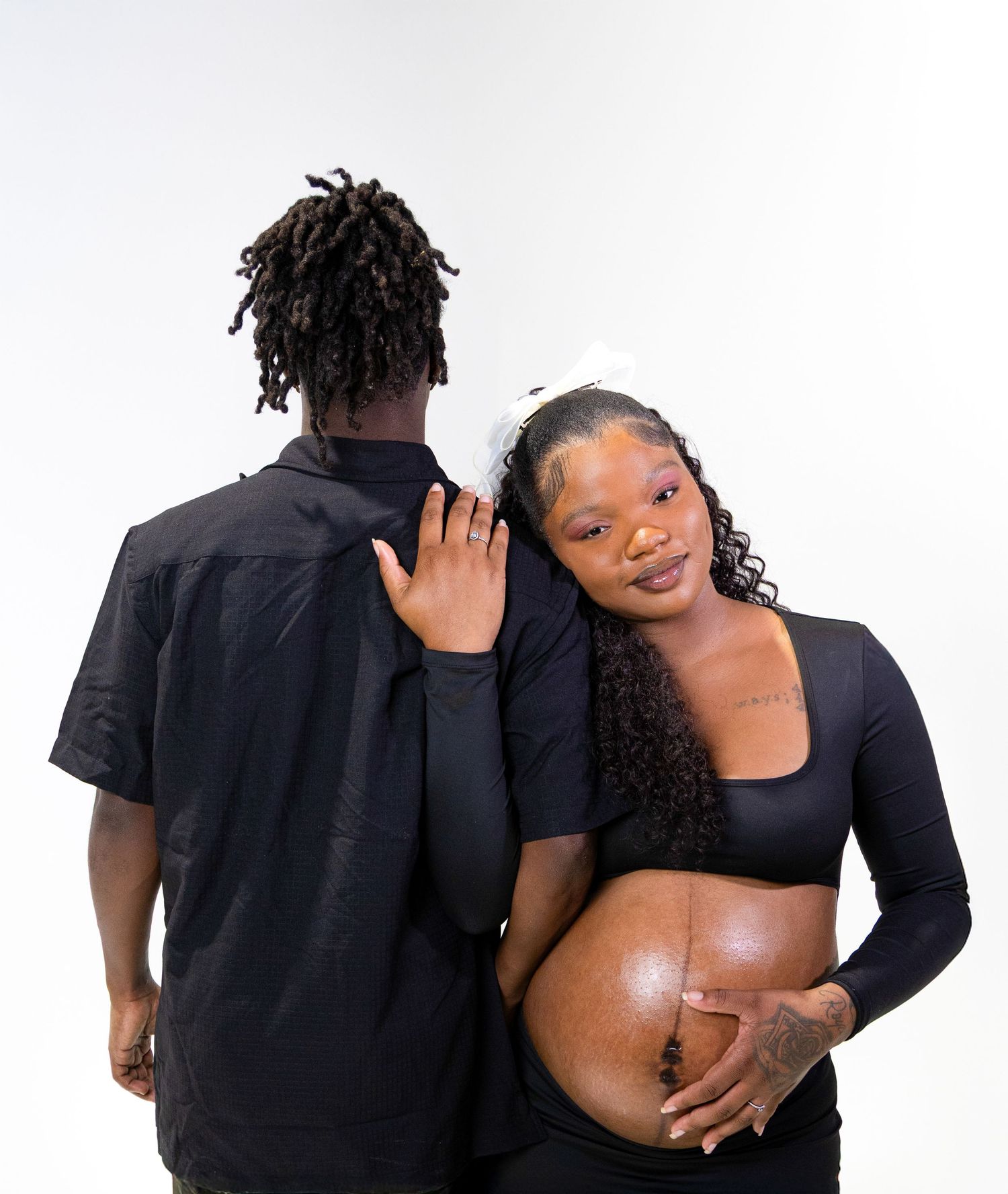 A pregnant woman in black maternity wear leans on her partner's shoulder, both dressed in black against a white backdrop.