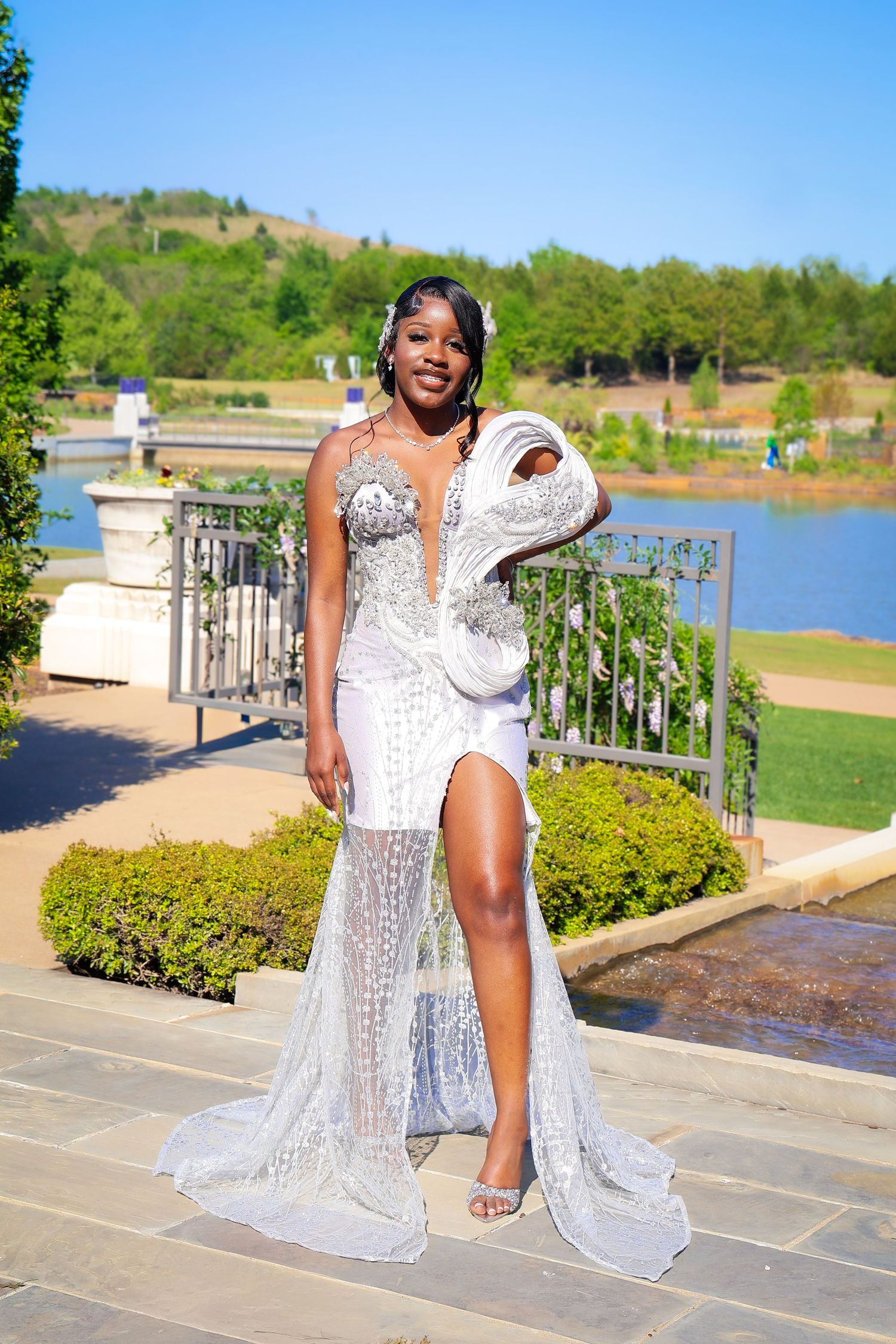Young Black woman in a sparkling white formal gown poses outdoors near a lake on a sunny day.