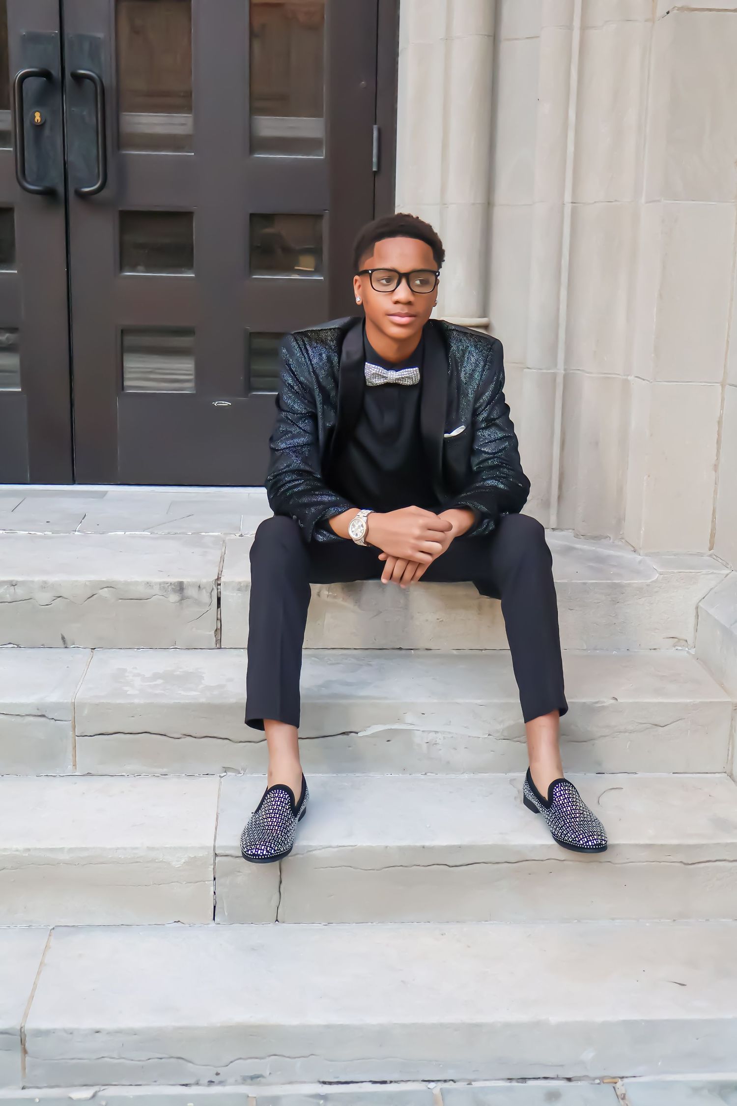 Dapper young man in black tuxedo and bow tie sitting on stone steps outdoors.
