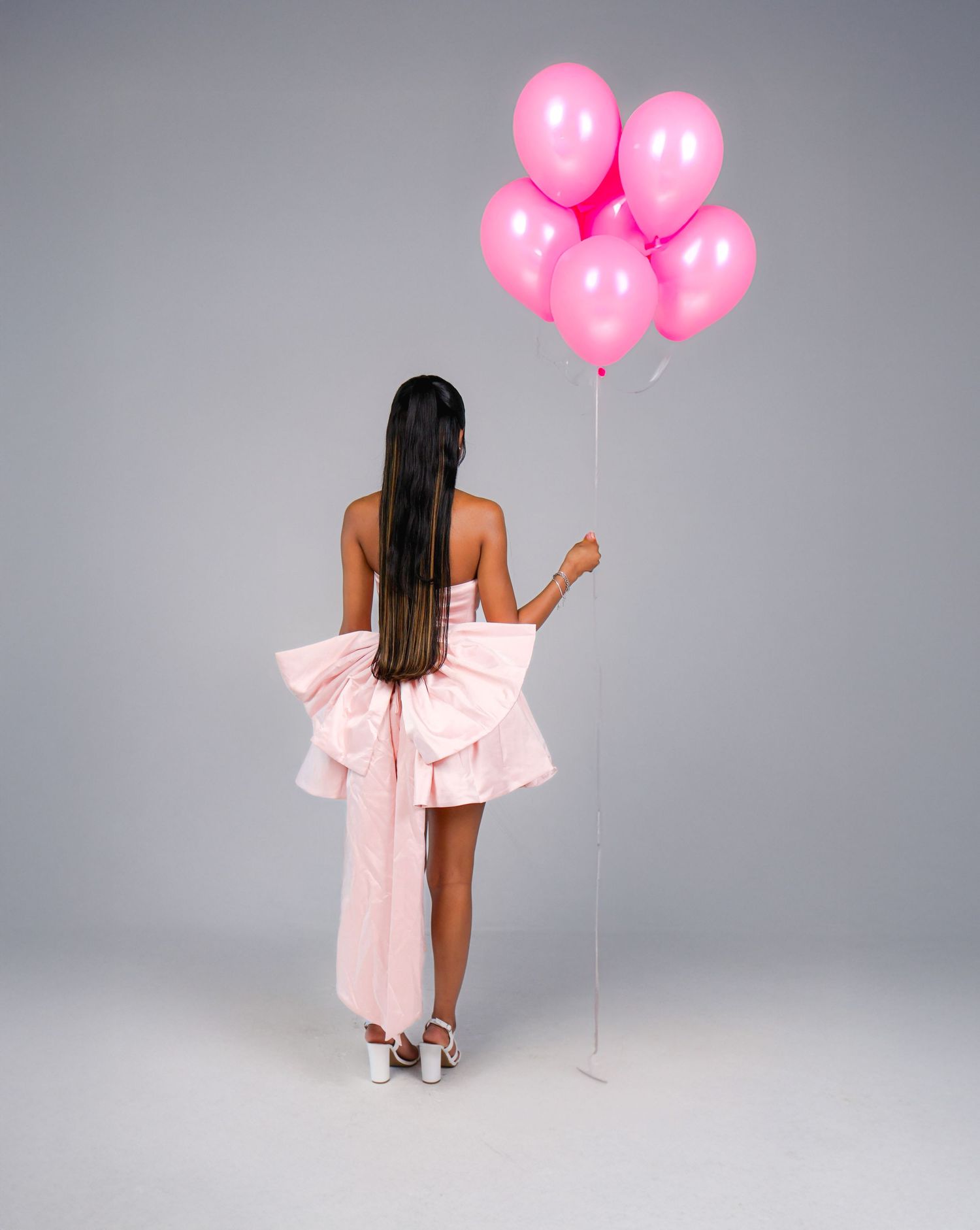 Woman in pink mini dress with bow holding pink balloons, back view, studio shot.