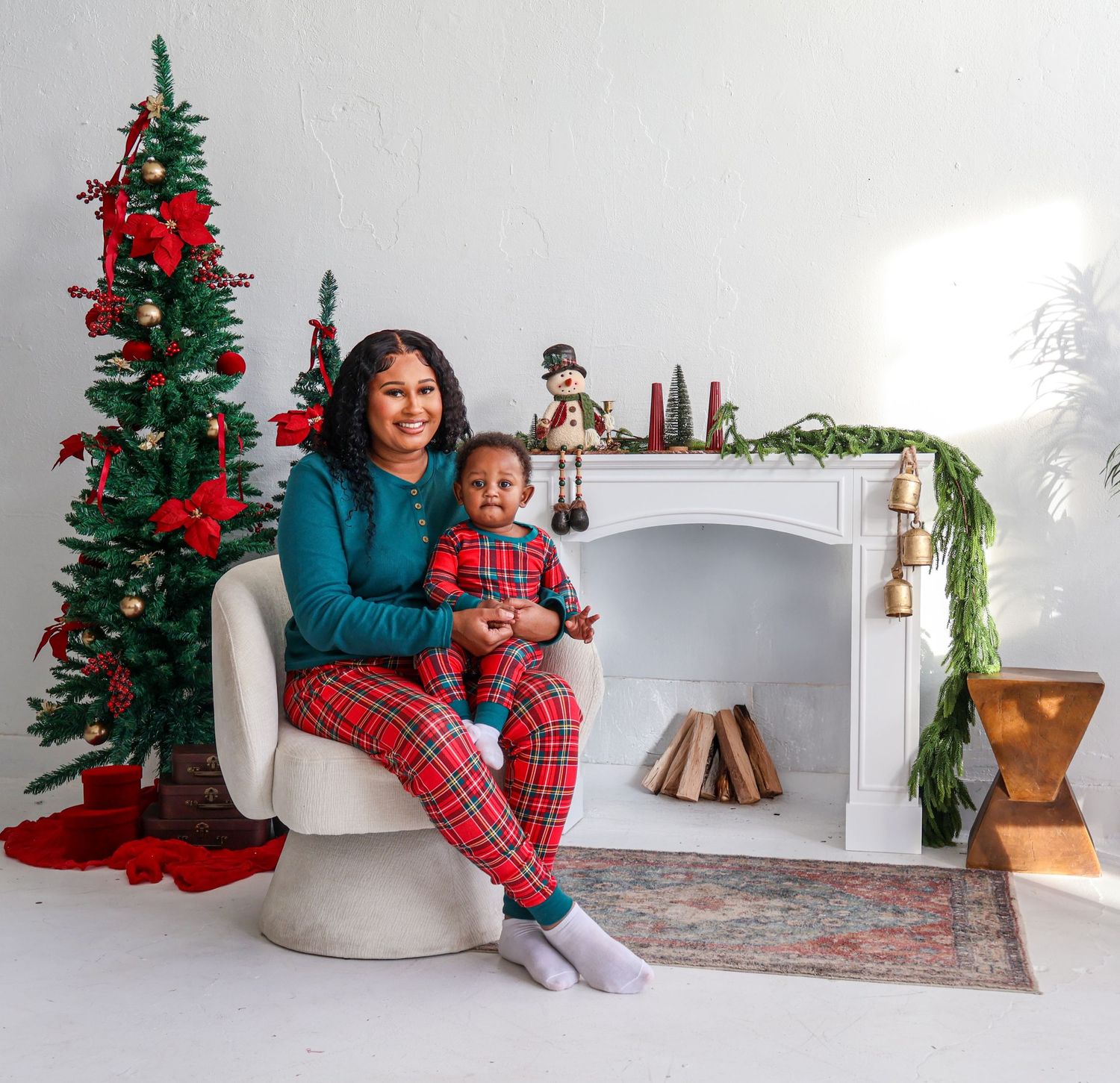 Mother and baby in matching red plaid Christmas pajamas sitting by decorated tree and fireplace.