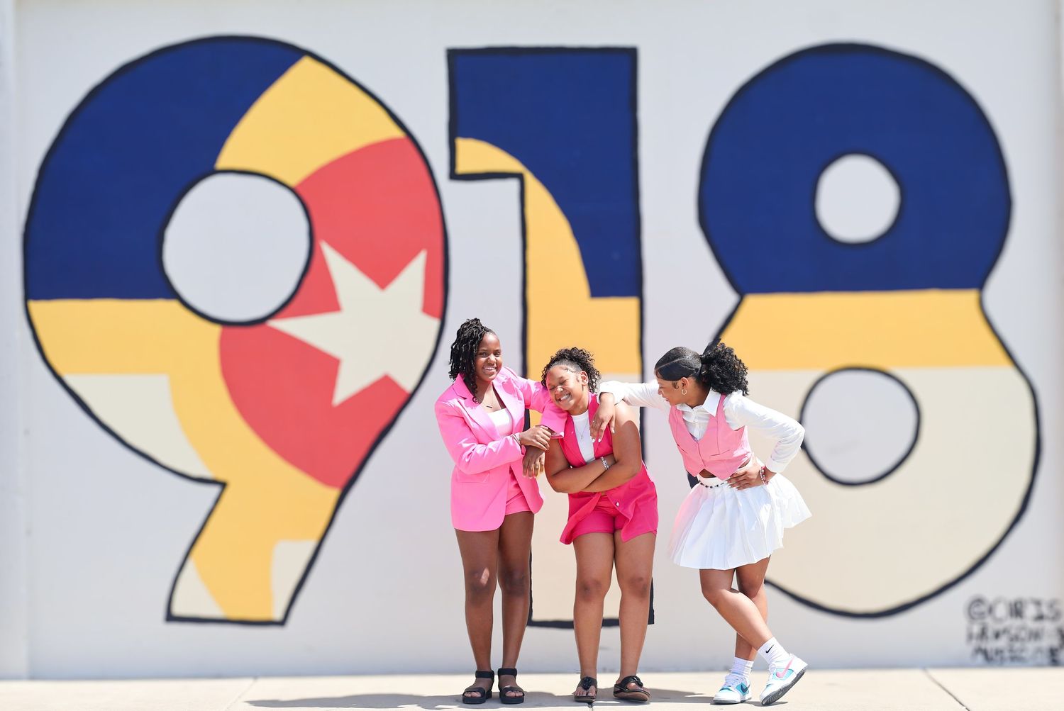 Three stylish women in pink outfits posing and laughing in front of a bold 918 street mural.